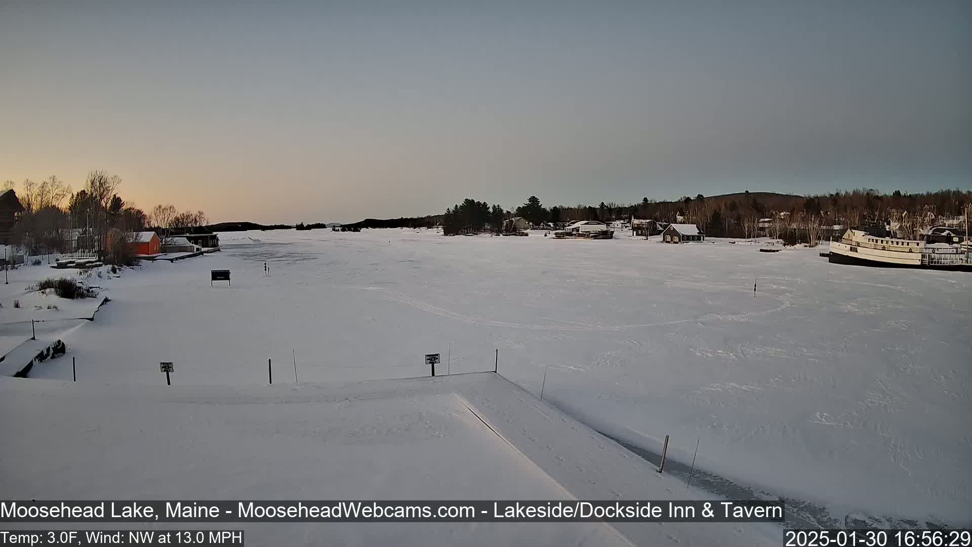 A frozen lake with a few buildings along the shore is visible under a twilight sky, with a northwest wind blowing at 13 mph.