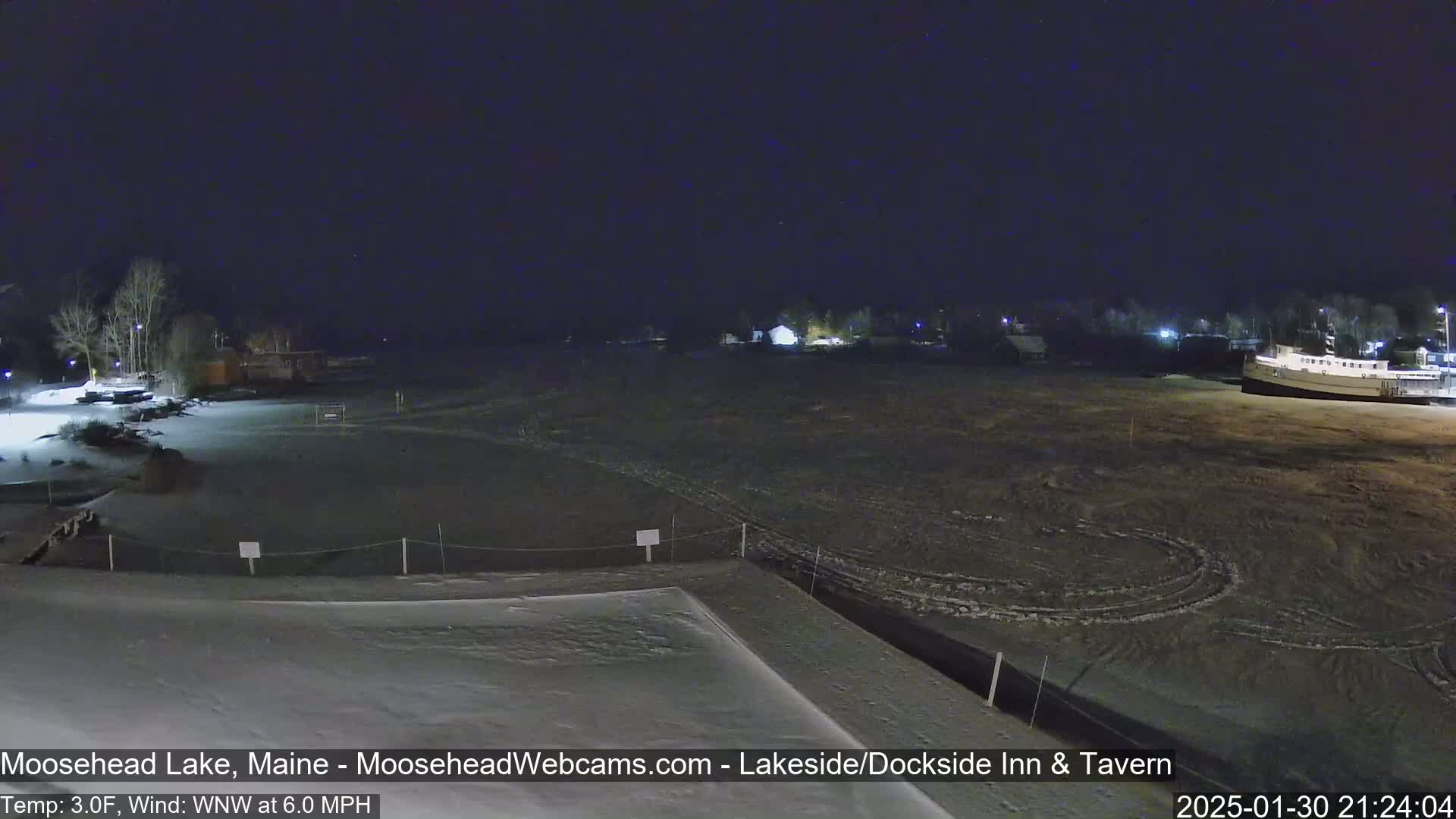 A nighttime, snow-covered lakeside scene with a few buildings and a boat visible under a clear, starry sky, with a temperature of 3°F and a wind blowing from the west-northwest at 6.0 mph.
