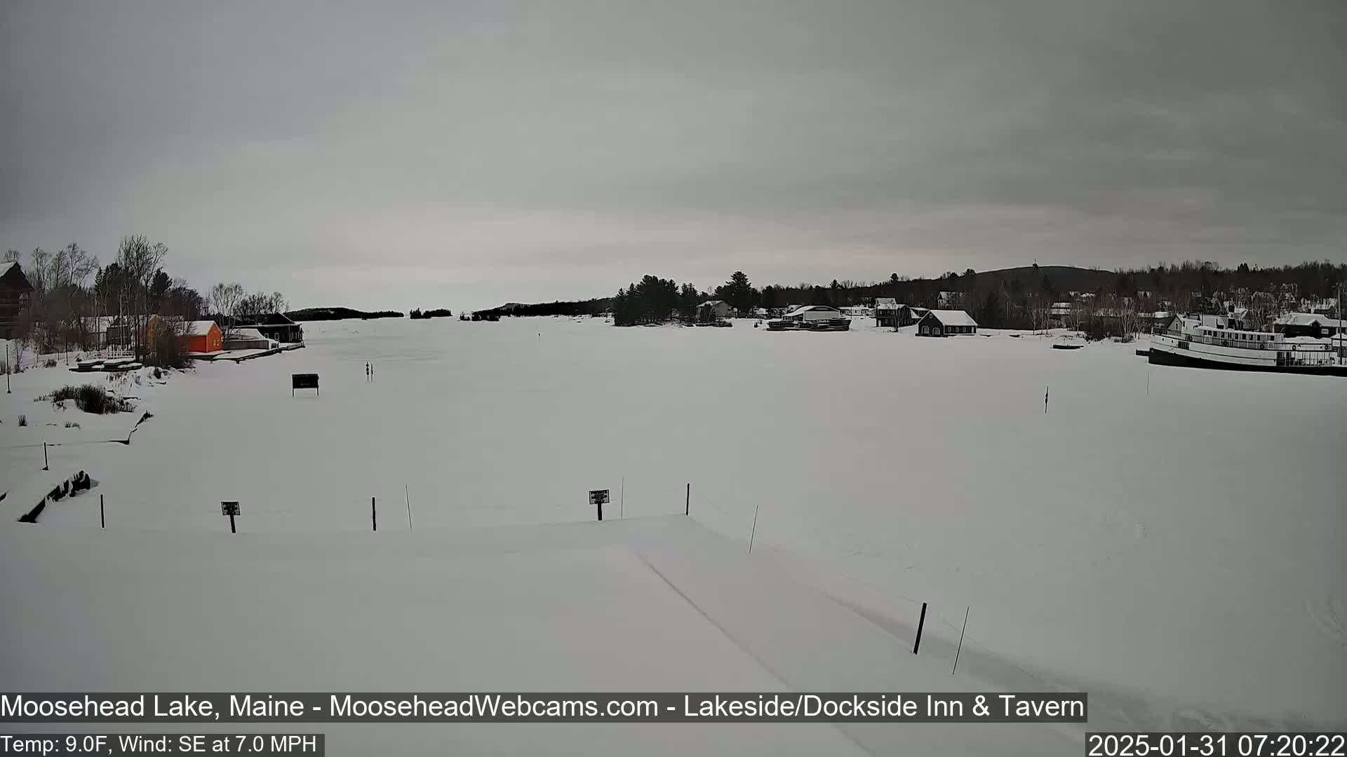 A snow-covered lake with buildings and a boat along the shore under an overcast sky.