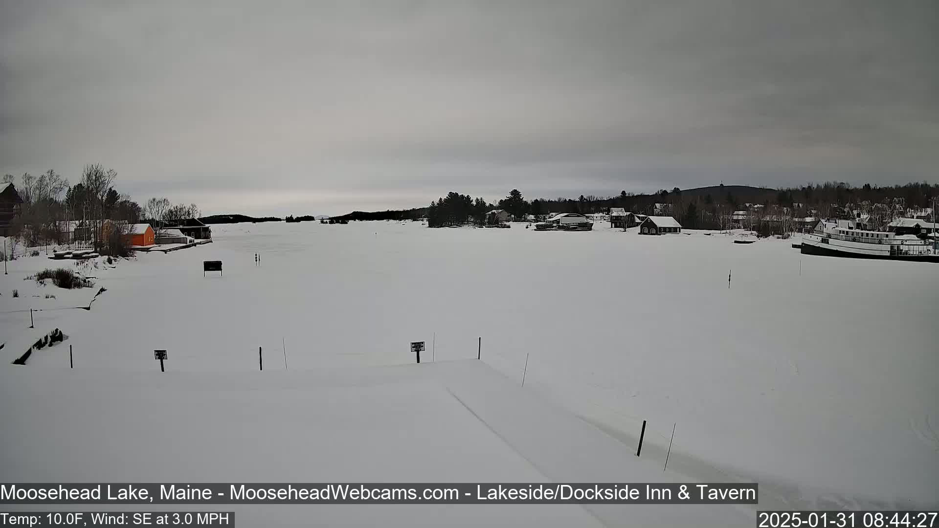 A snow-covered lake with buildings and a boat visible in the distance under an overcast sky.