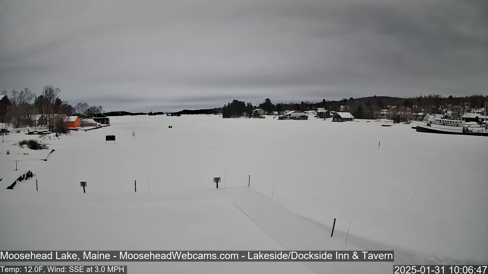 A snow-covered lake with buildings and a boat in the distance under an overcast sky.