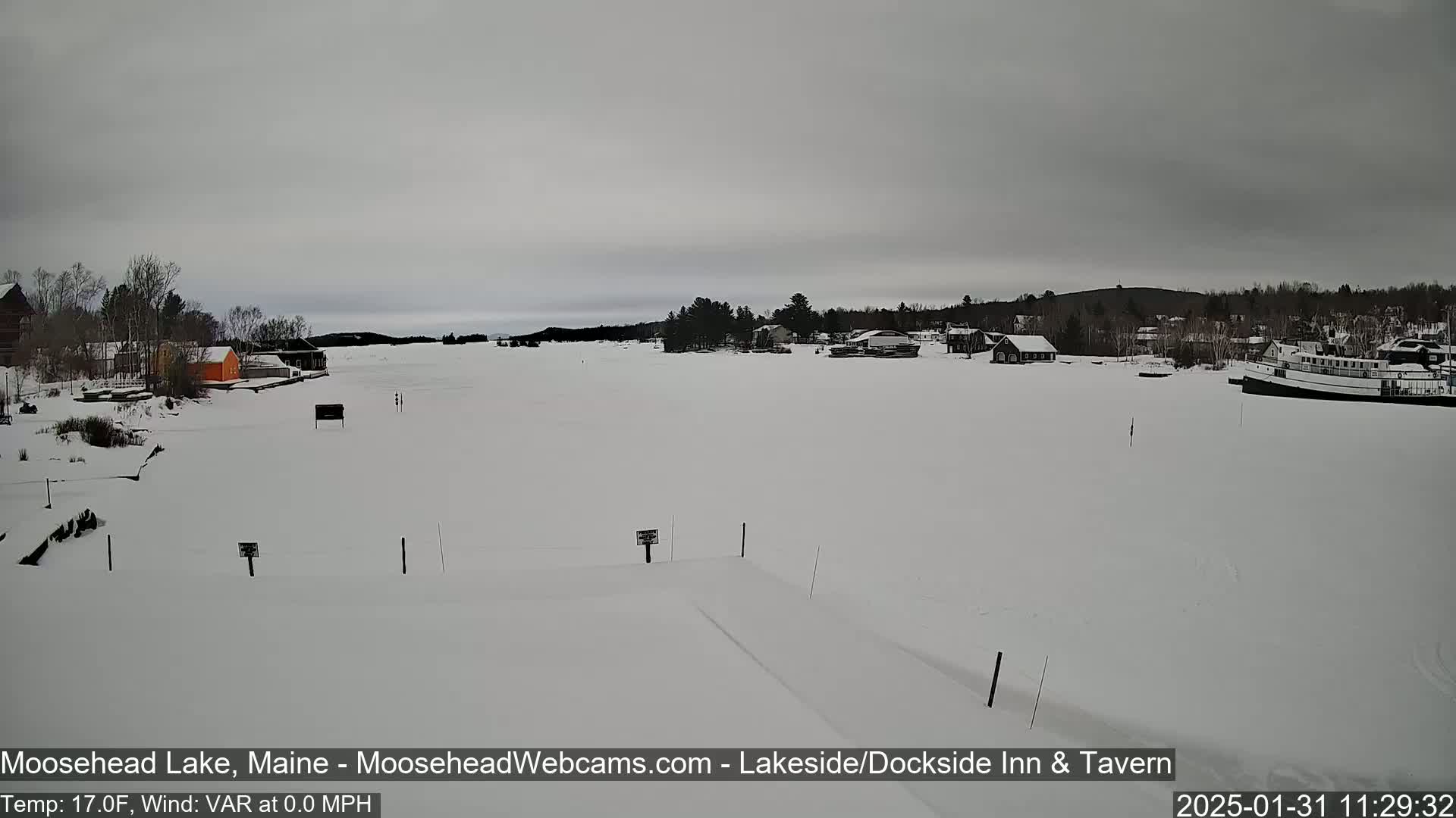 A snow-covered lake with a few buildings and a boat visible under an overcast sky.