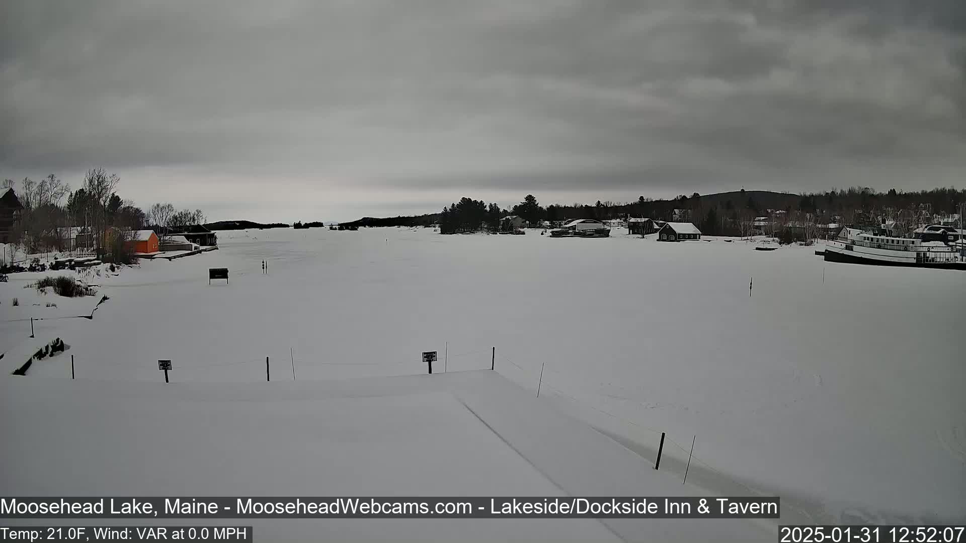 A snow-covered frozen lake is seen with a few buildings and a boat visible in the distance under an overcast sky.
