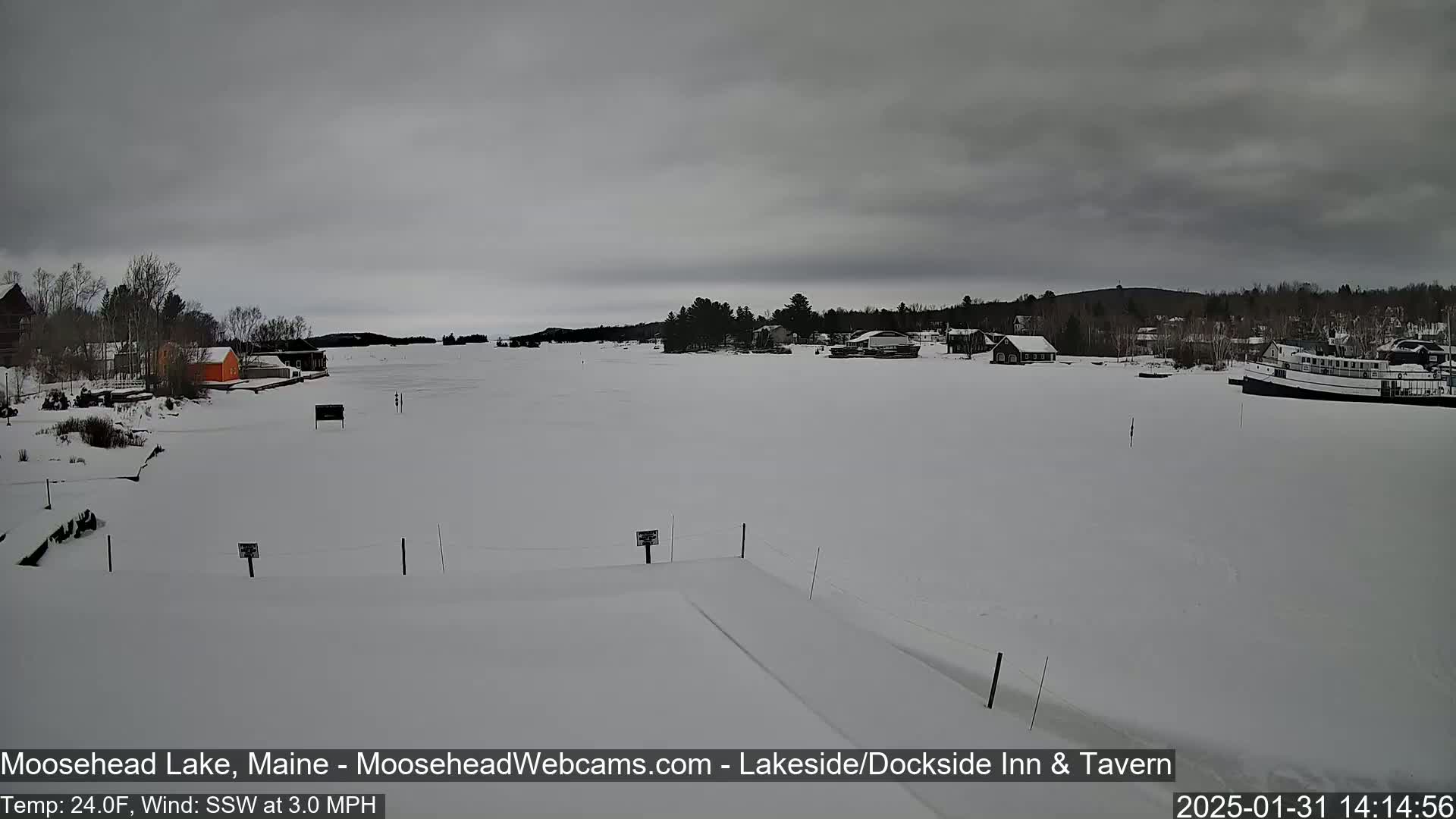 A snow-covered frozen lake is seen under an overcast sky, with a few buildings and a boat visible in the distance.