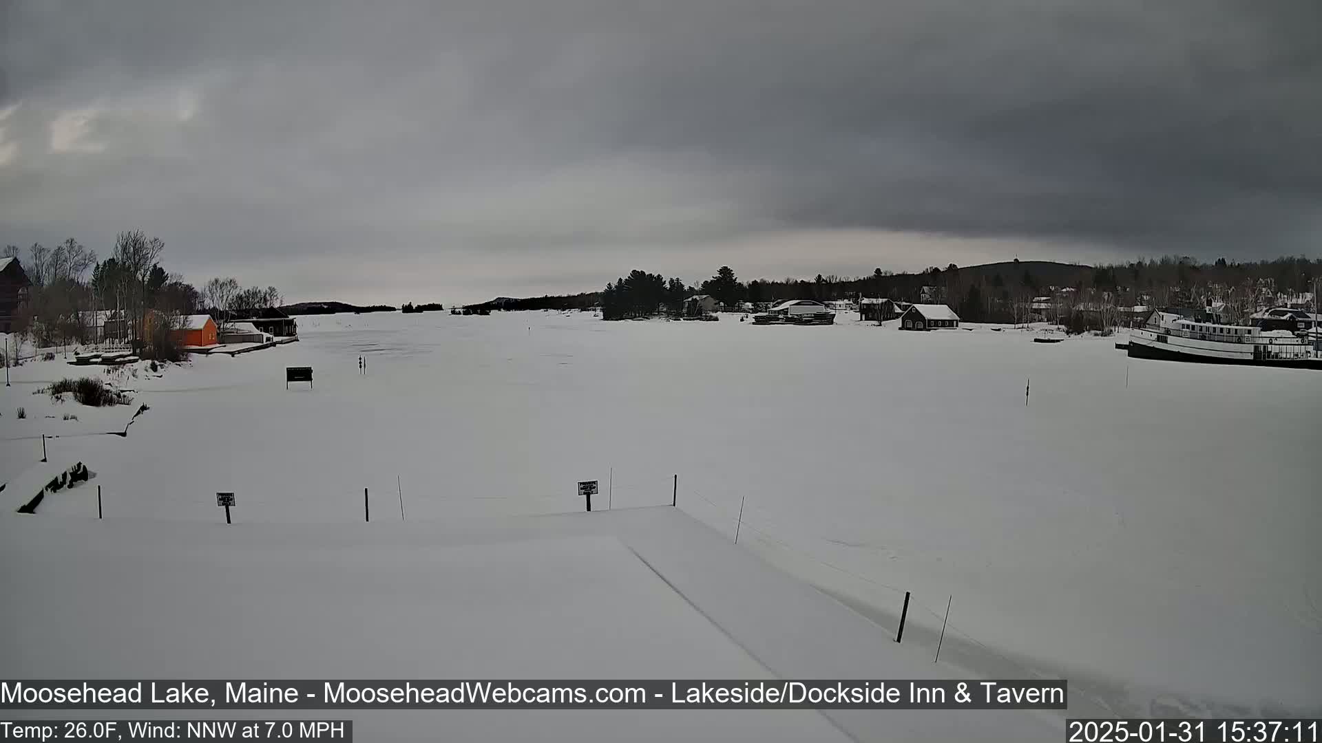 A snow-covered frozen lake is seen under an overcast sky, with buildings and trees visible along its edge.