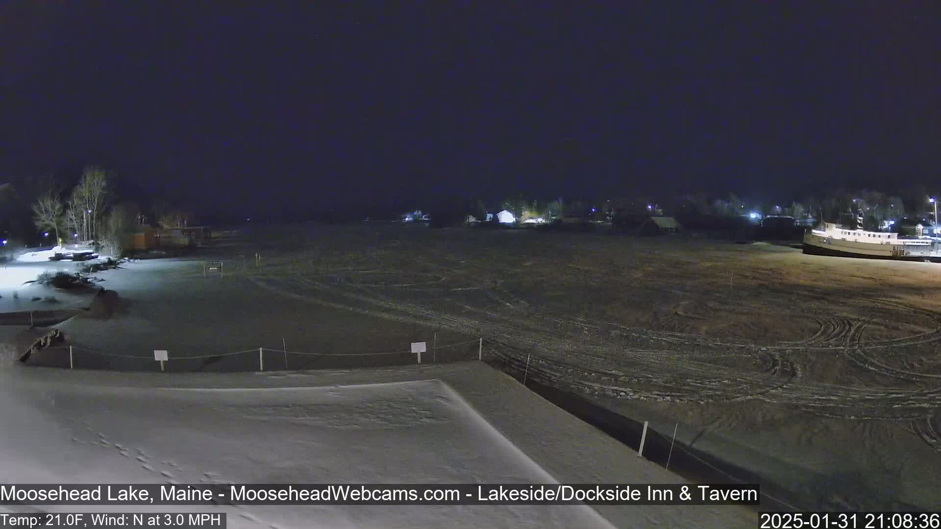 A snow-covered lakeside area at night, with a few buildings visible in the distance under a dark sky, a north wind blowing at 3 mph, and the temperature at 21°F.