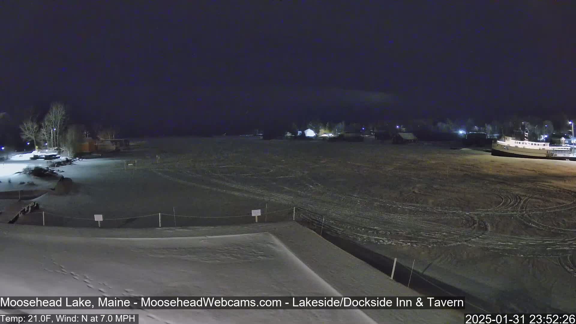 A nighttime view of a snow-covered lakefront scene with a few buildings and trees visible under a dark sky, with a north wind at 7 mph and a temperature of 21°F.