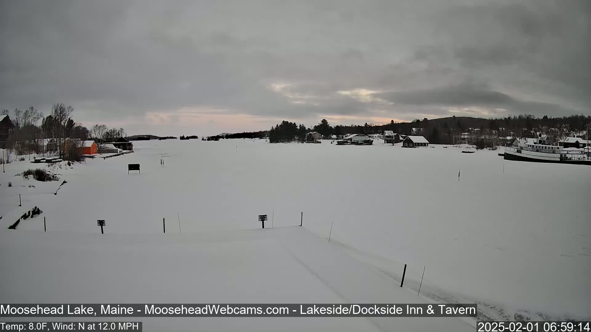 A snow-covered lake with a few buildings and a boat on the far shore under a cloudy, overcast sky.