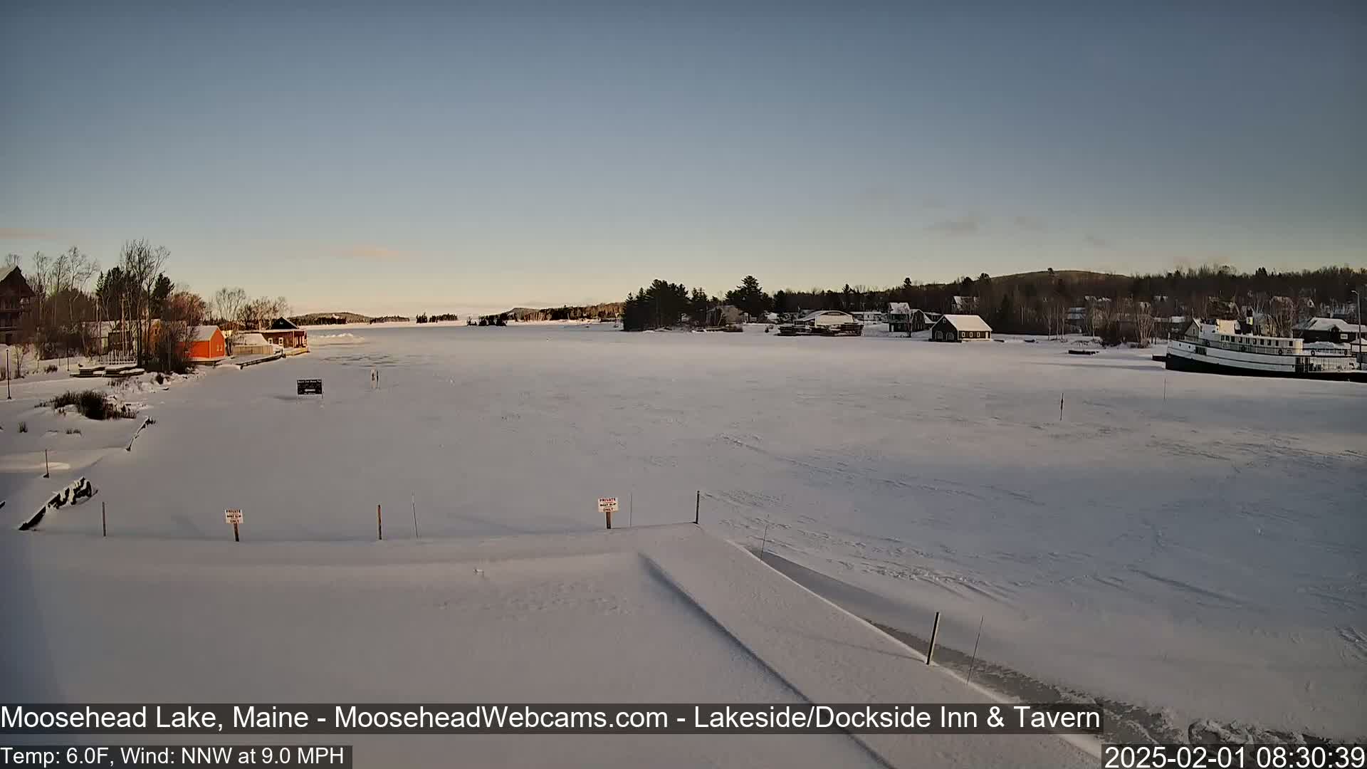 A frozen lake, under a clear sky, features buildings along the shoreline and a boat moored to the ice.