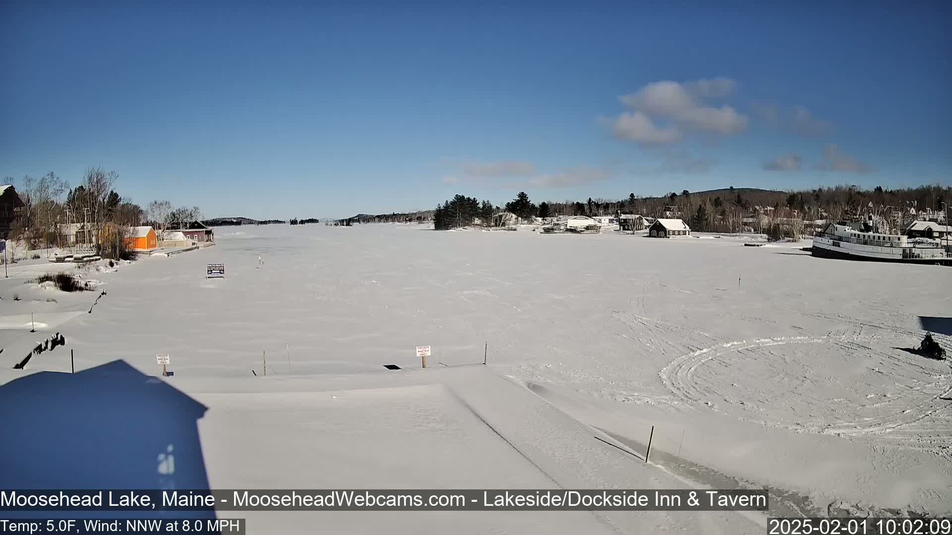 A snow-covered frozen lake with a few buildings and a boat visible on a sunny day with a light northwest wind.