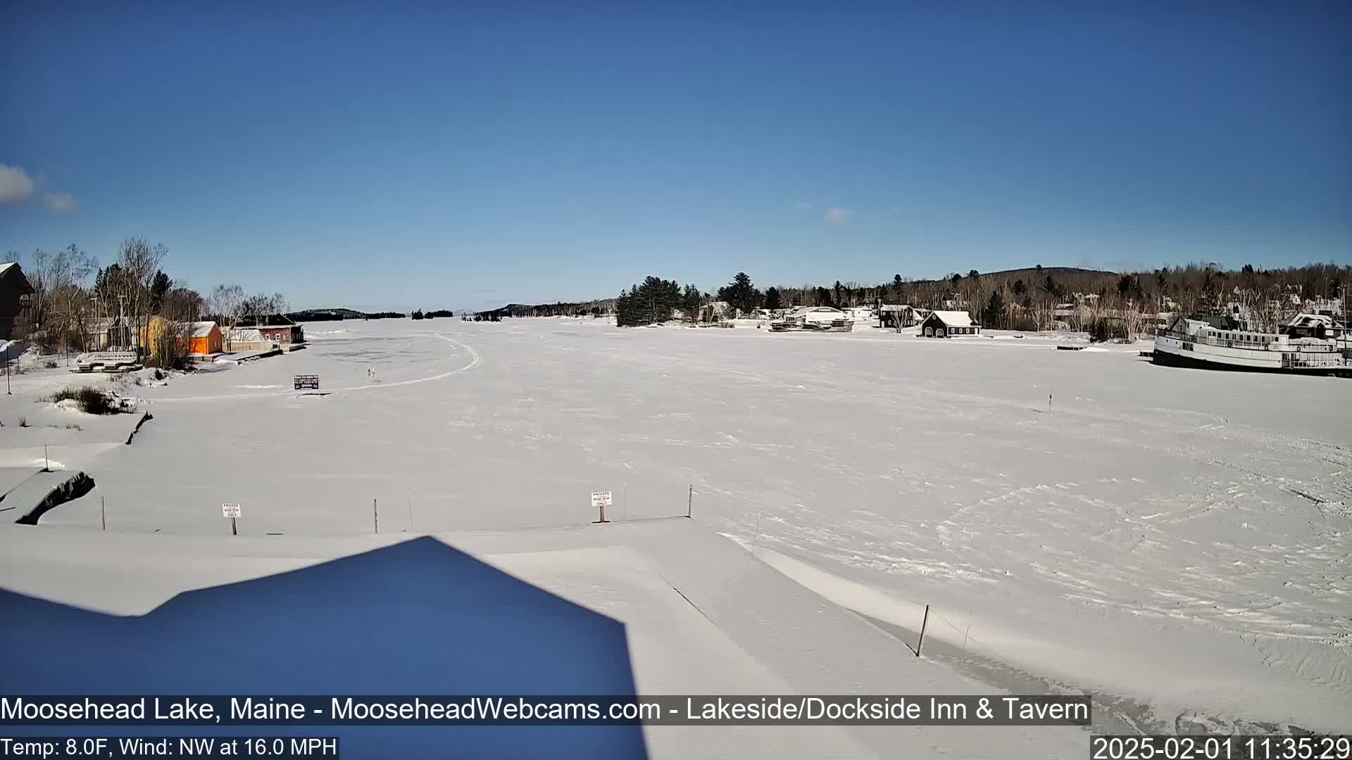 A frozen lake on a sunny day with a northwest wind, showing buildings and a boat along the shoreline.