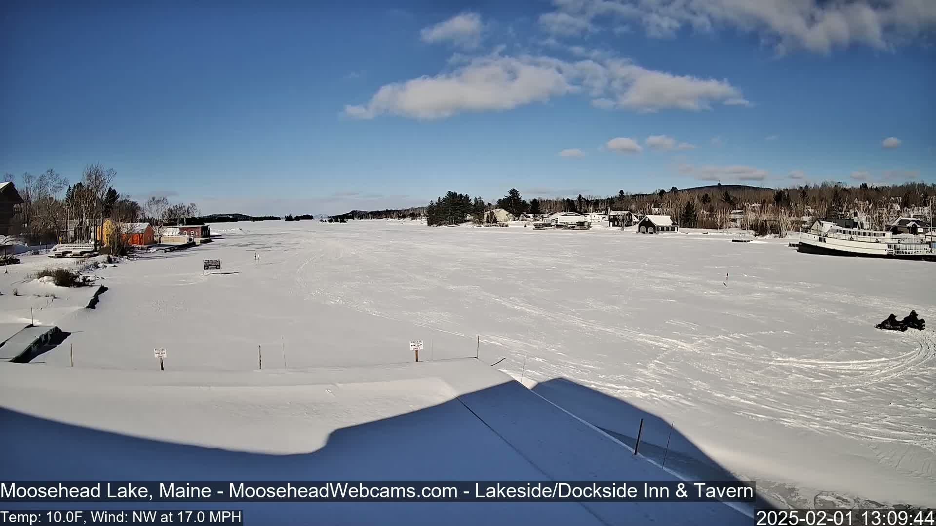 A snow-covered frozen lake with buildings and trees on the shore under a mostly sunny sky.