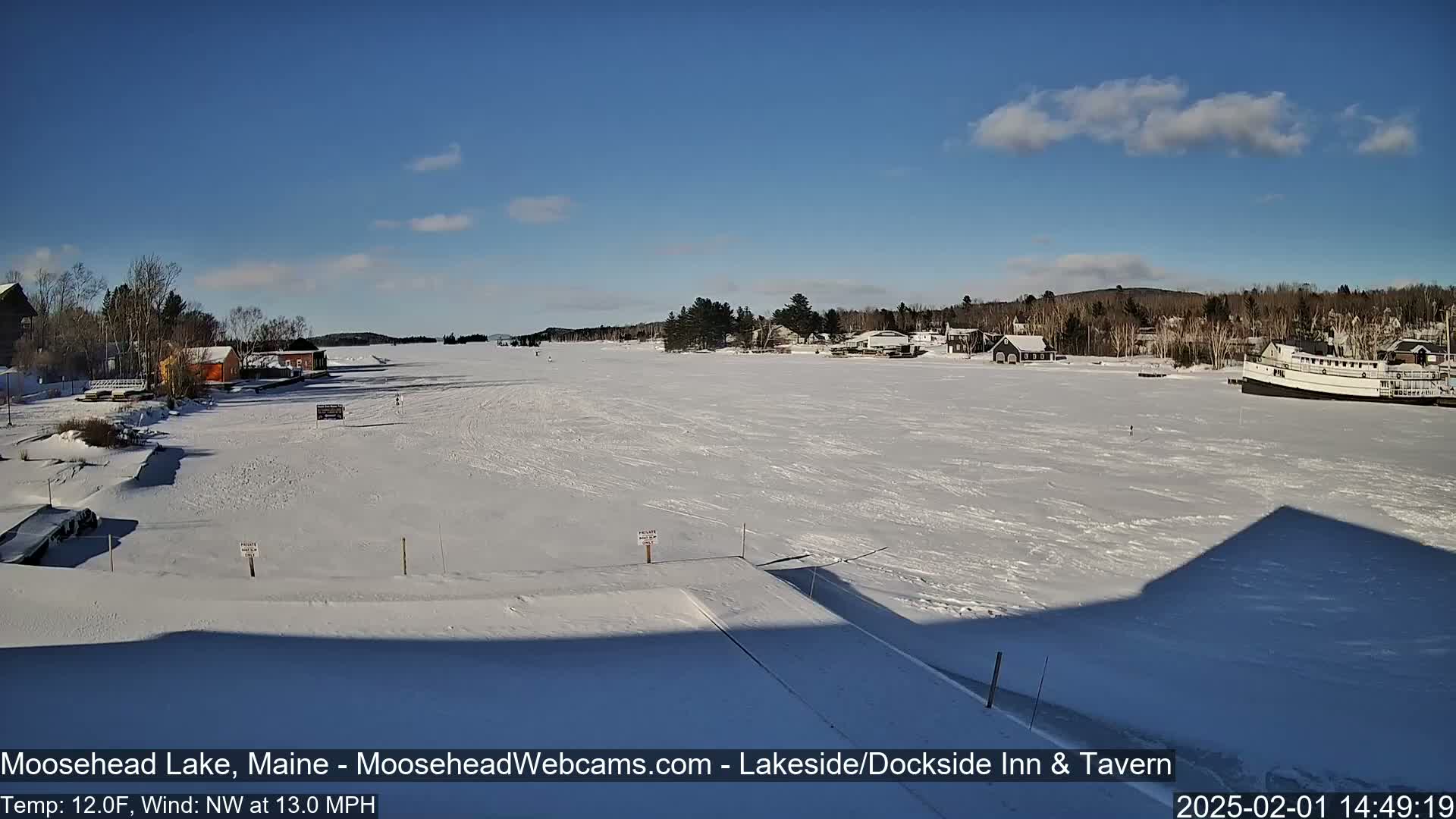A frozen lake under a mostly clear, sunny sky shows a few buildings and a boat on the far shore.