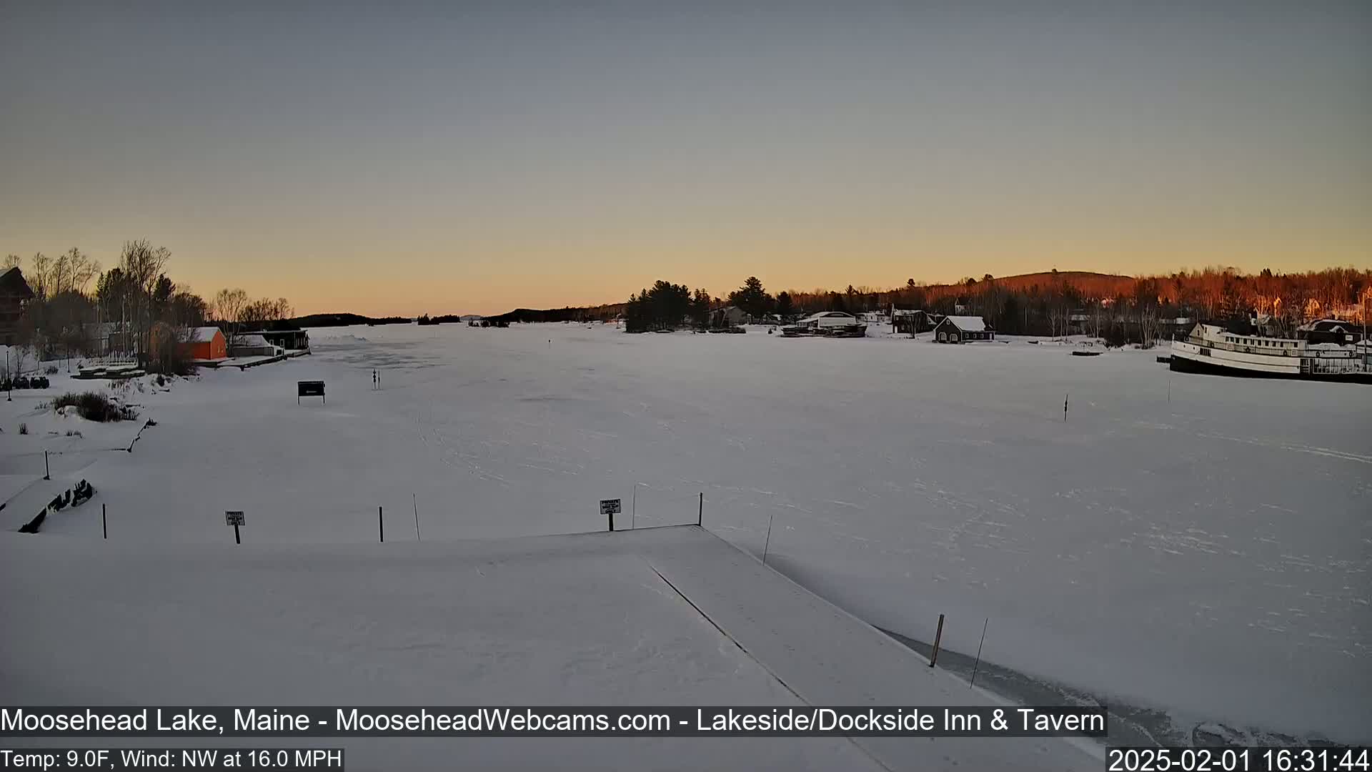 A frozen lake at sunset, with a few buildings and a boat visible on the shore under a clear sky, and a northwest wind blowing at 16 mph.