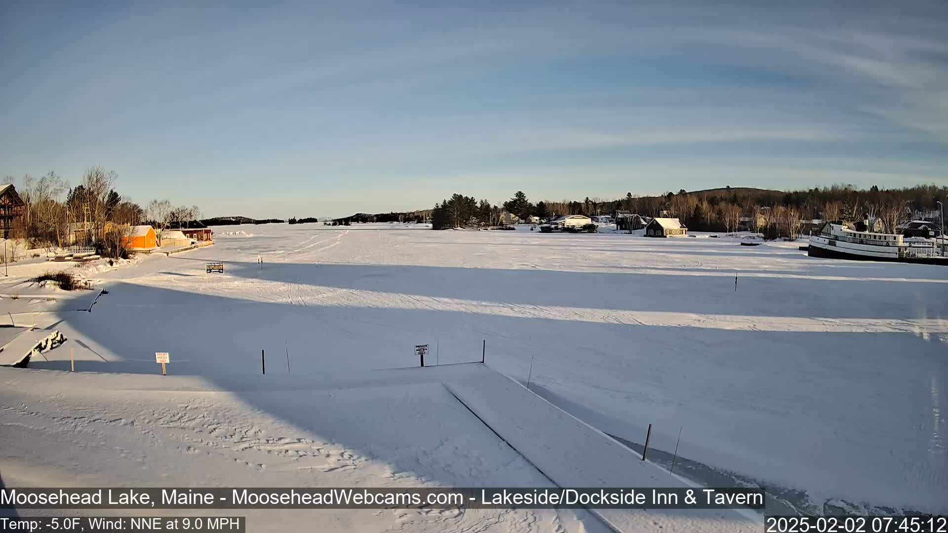 A frozen lake is seen on a sunny, cold day, with a few buildings and a boat visible in the distance.