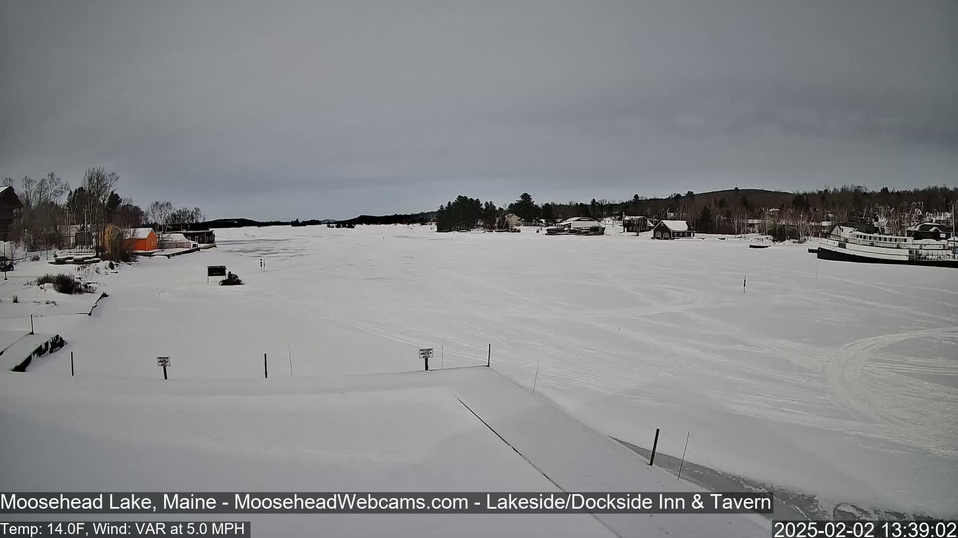 A snow-covered frozen lake with several buildings and a boat on the far shore under an overcast sky.