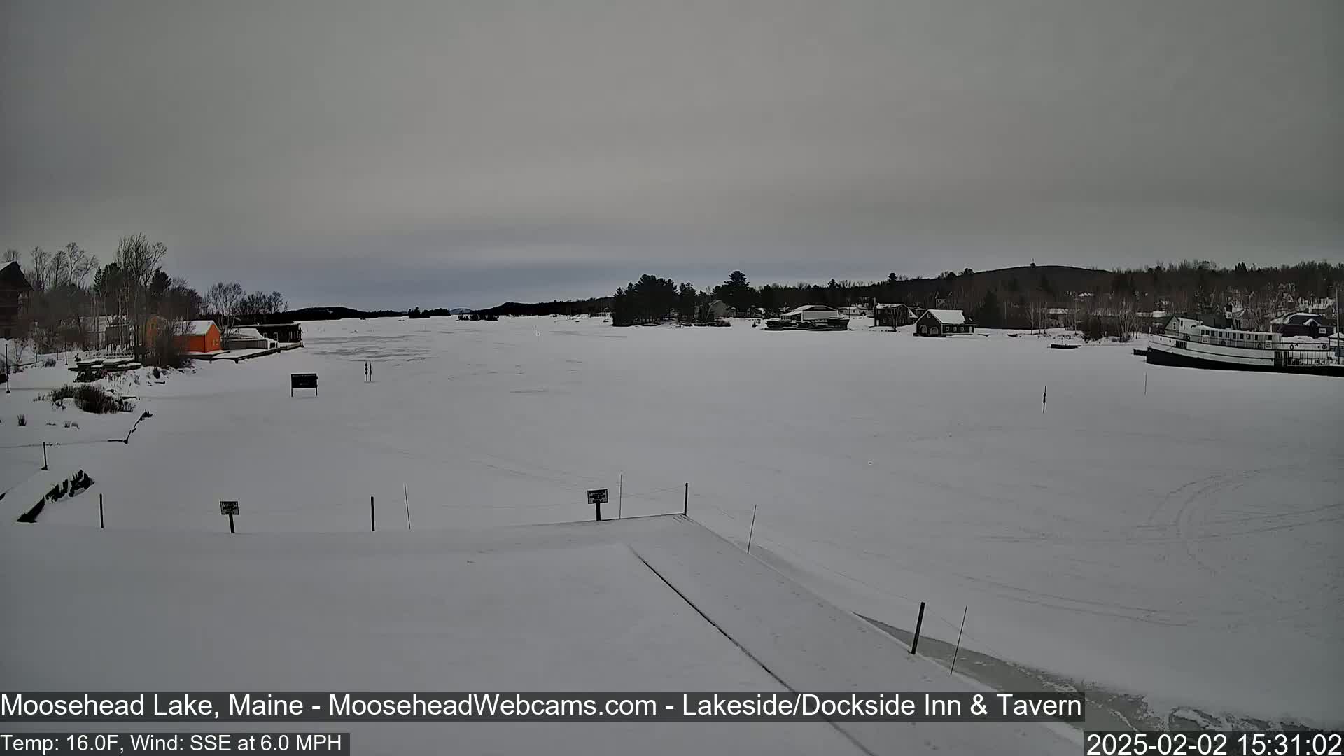 A frozen lake is visible, under an overcast sky, with some buildings and a boat docked on the shore.