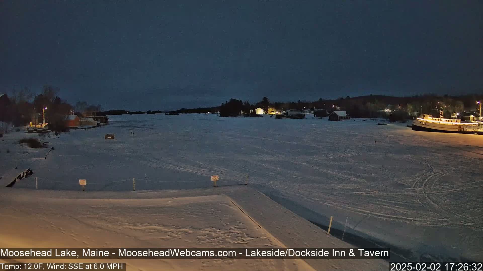 A snow-covered frozen lake at night, with a few lights visible on the far shore and a large boat docked near the right edge.