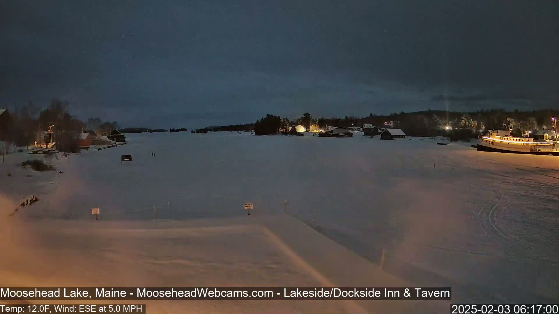 A frozen lake at night, under a dark sky, shows a few buildings with lights on along the shore and a boat docked on the ice.