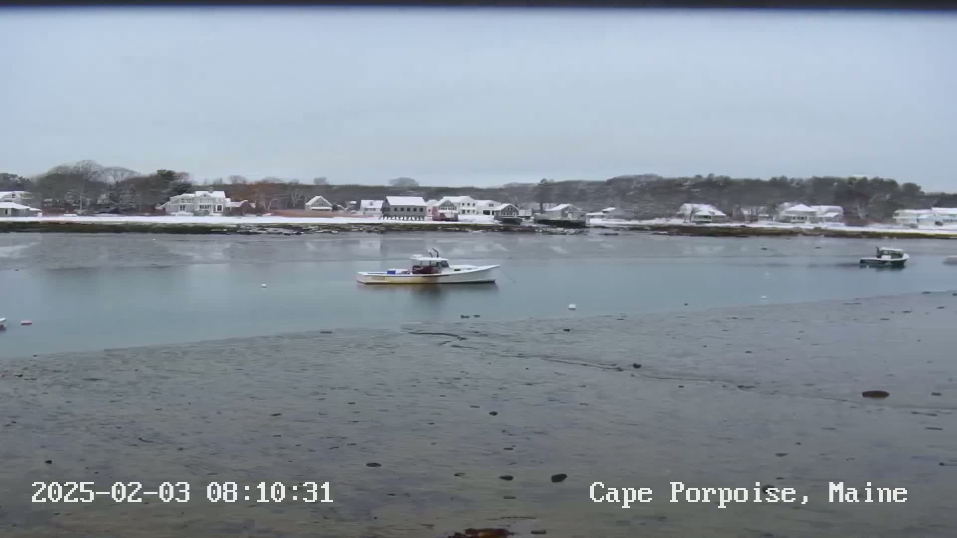 A snow-covered waterfront town is visible across a calm body of water where several small boats are anchored; the foreground shows exposed mudflats.