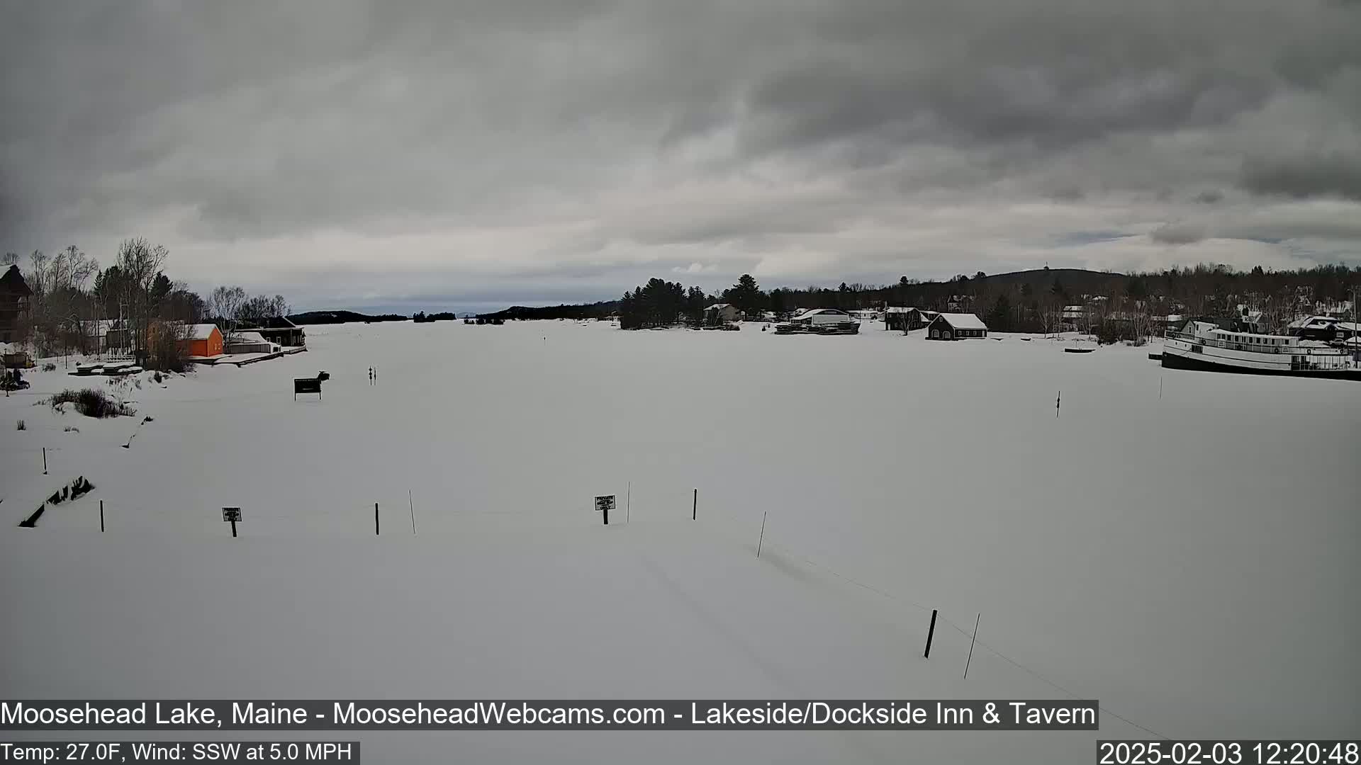 A frozen lake, under an overcast sky, shows a few buildings and a boat in the distance.