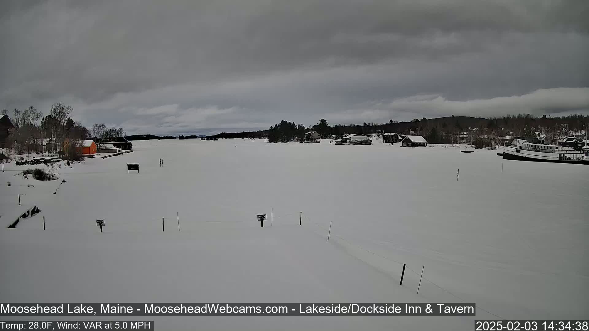 A snow-covered lake with buildings and trees along the shore under a cloudy sky.