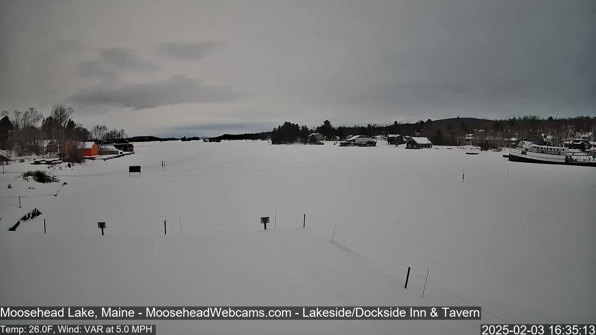 A snow-covered frozen lake is seen under an overcast sky, with a few buildings and a boat visible in the distance.