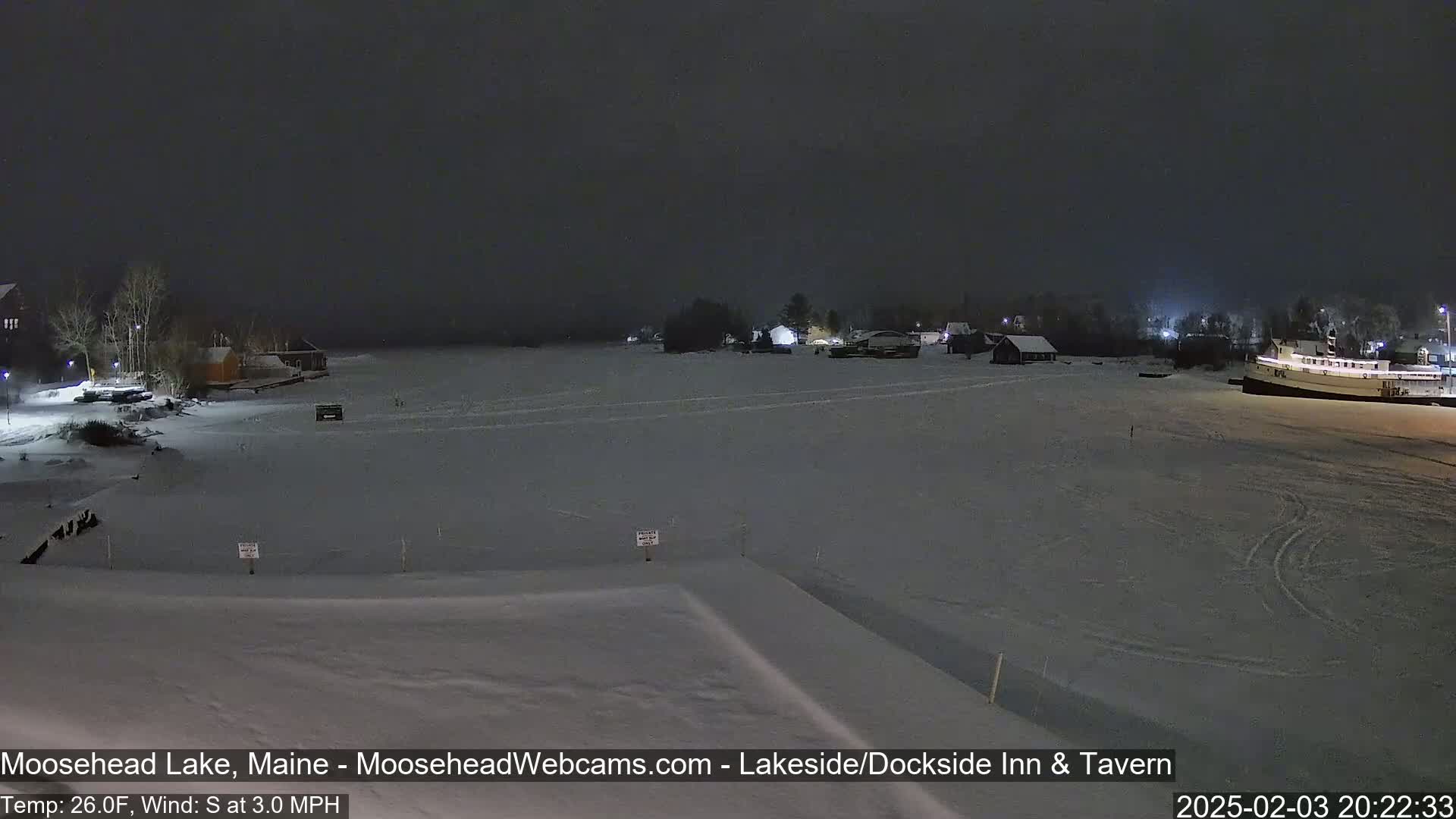A nighttime view of a snow-covered frozen lake with buildings and a boat visible under a dark, cloudy sky.