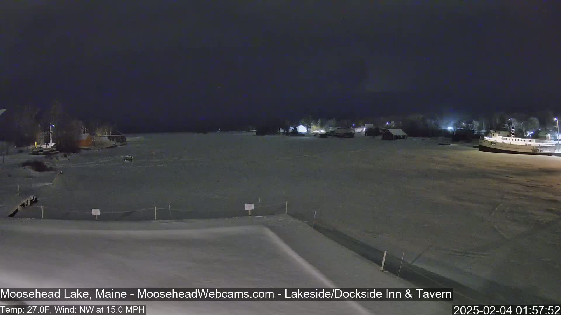 A nighttime view of a snow-covered lake with buildings and a boat in the distance, under a dark sky with a northwest wind of 15 mph and a temperature of 27°F.