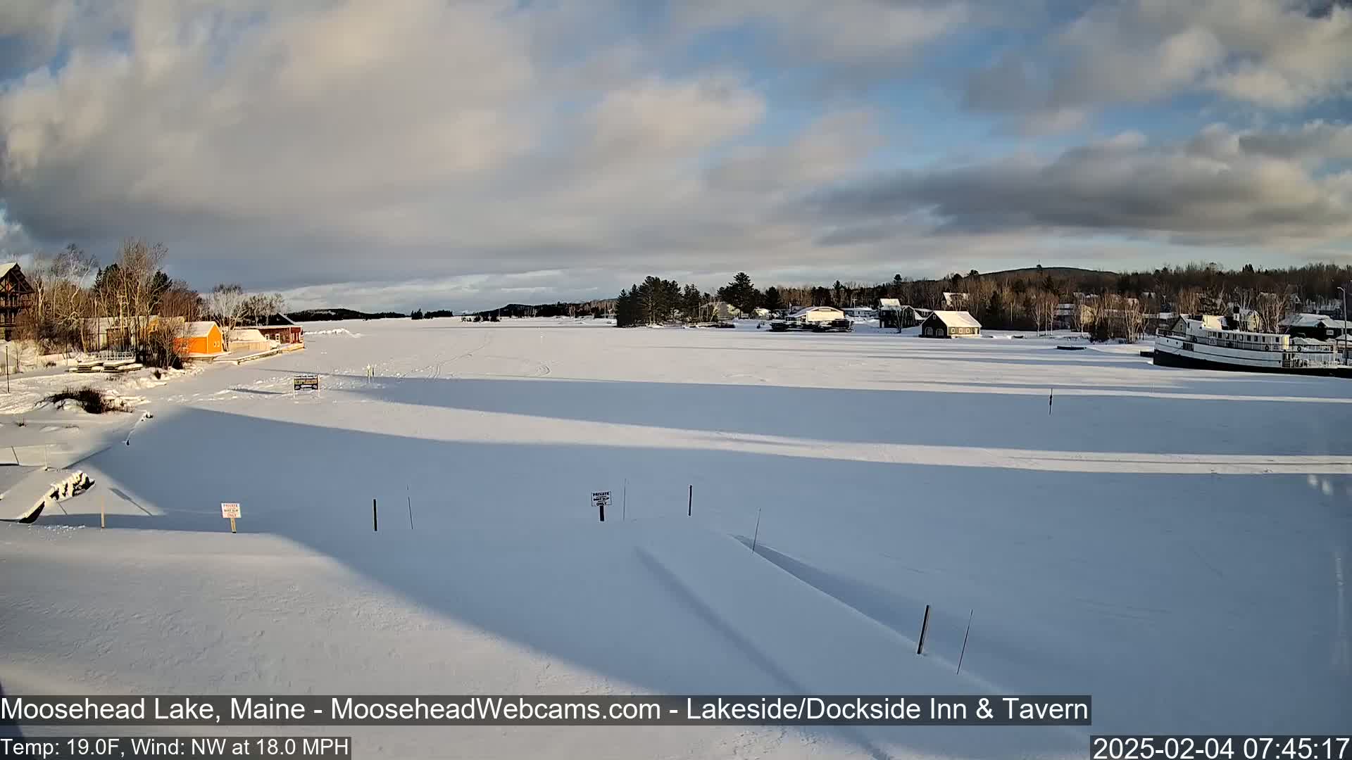 A frozen lake is seen under a partly cloudy sky, with buildings and a boat visible in the distance, and snow covering the ground in the foreground.