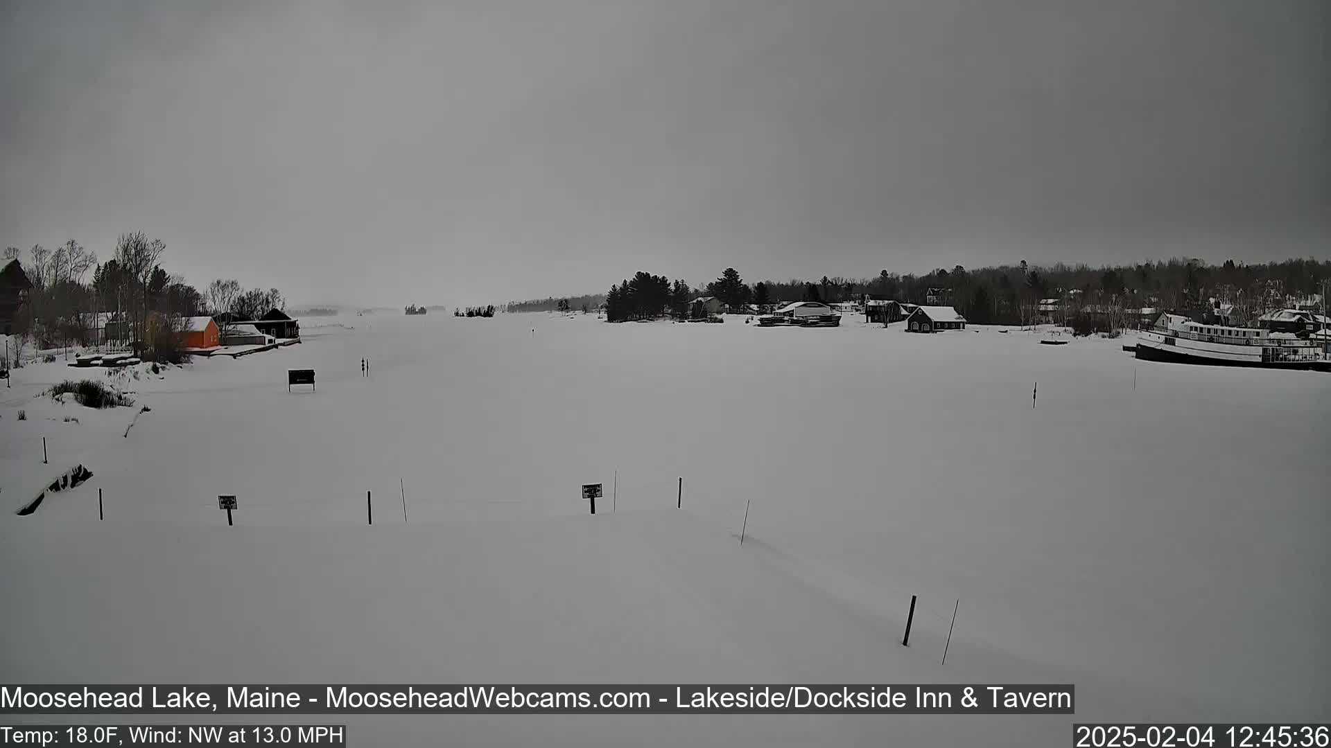 A snow-covered frozen lake is seen with a few buildings and a boat in the distance under an overcast sky.
