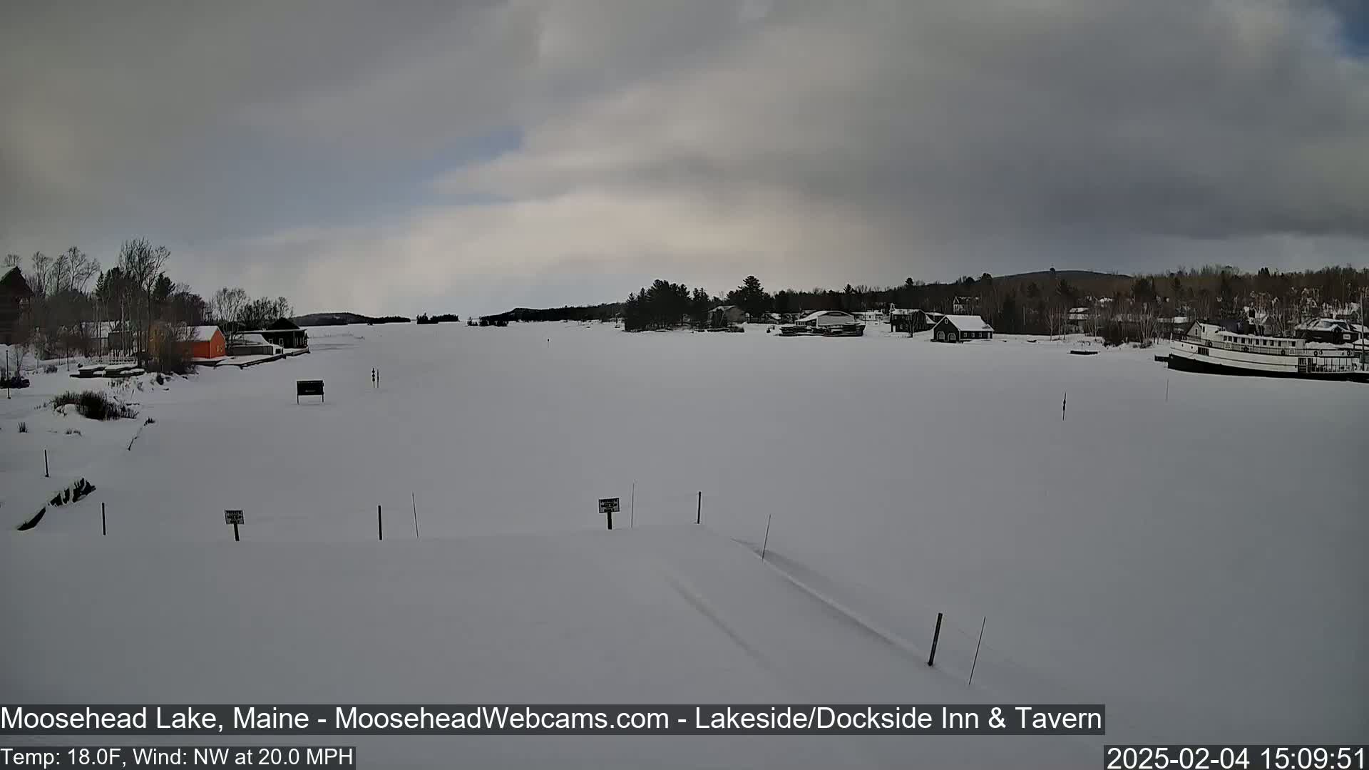 A snow-covered frozen lake stretches out before a shoreline with buildings and trees under an overcast sky.