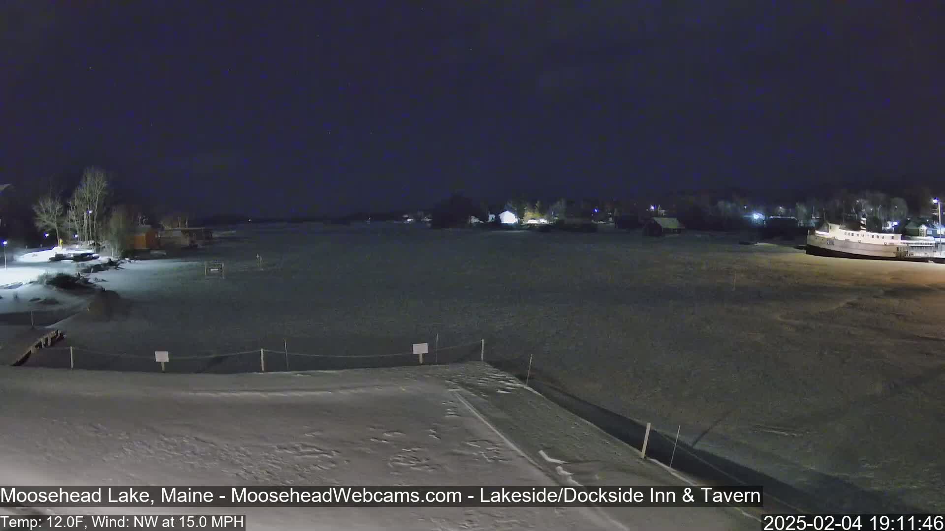 A snow-covered lakeshore at night, with a few buildings and a boat visible in the distance under a dark sky.