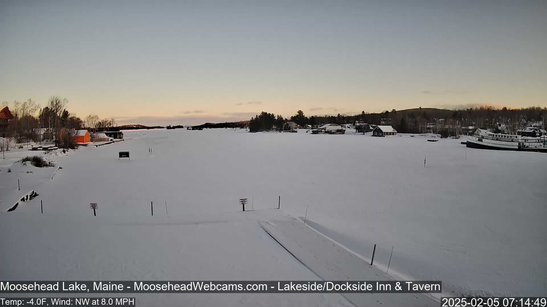 A frozen lake at sunset, with a few buildings and a boat visible on the shore, under a mostly clear sky with a temperature of -4°F and a northwest wind at 8 mph.