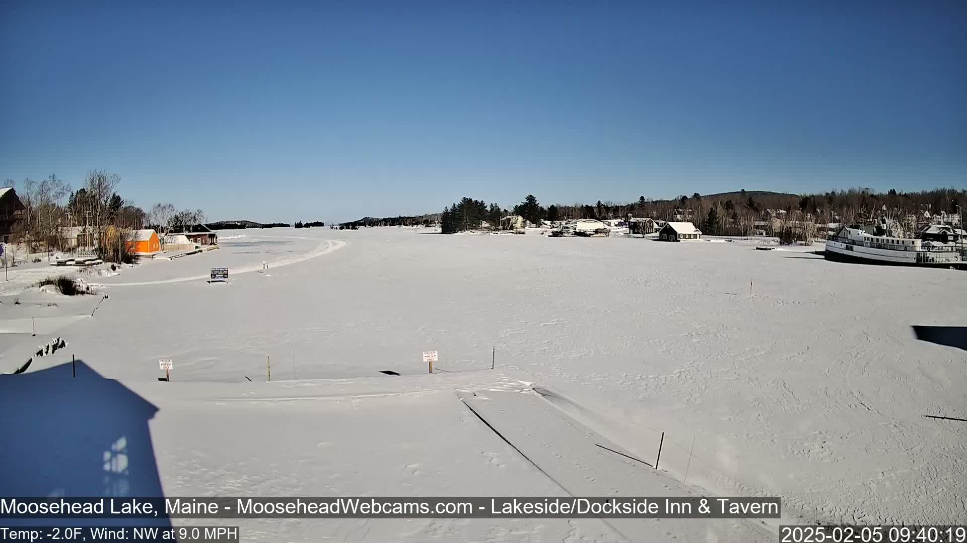 A frozen lake, under a clear blue sky, features a snow-covered shoreline with buildings and a docked boat.