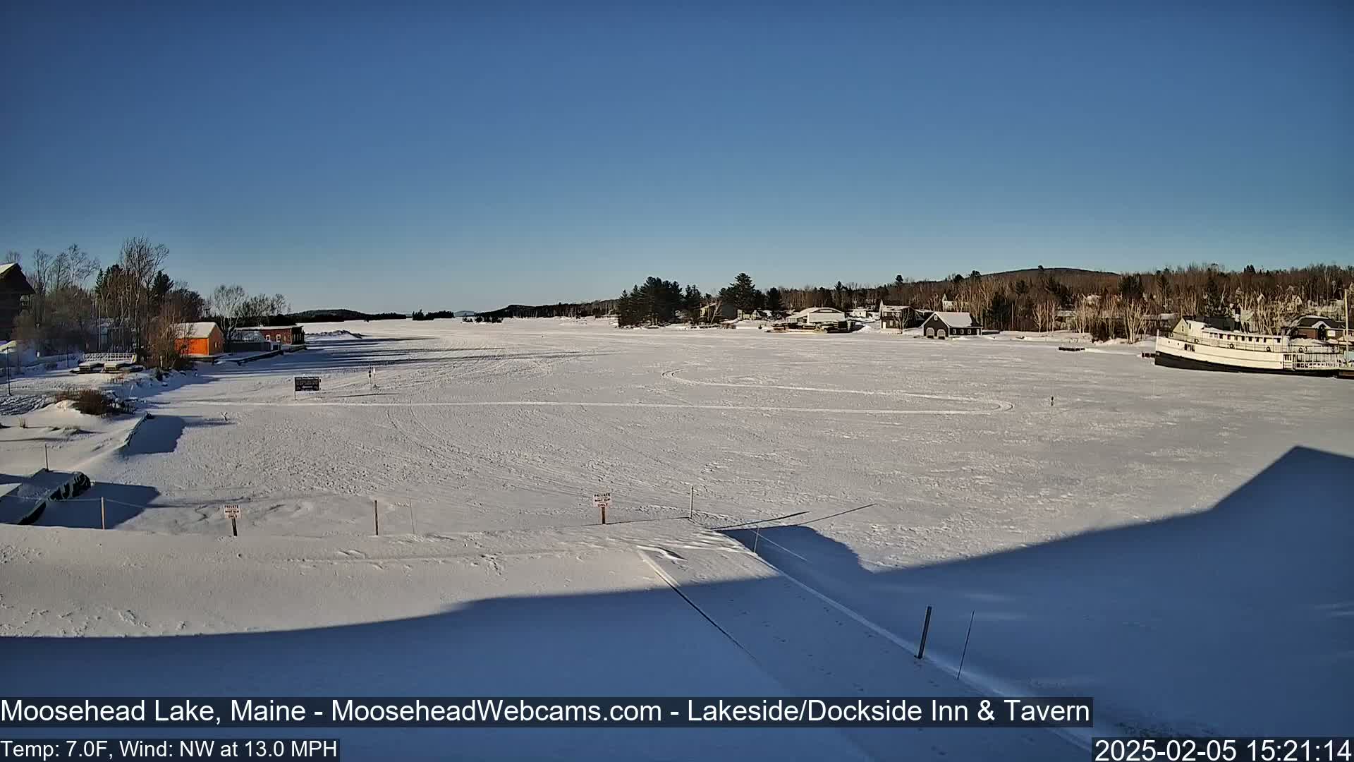 A frozen lake under a clear blue sky, with a few buildings and a boat visible along the shoreline, and a northwest wind blowing at 13 mph.