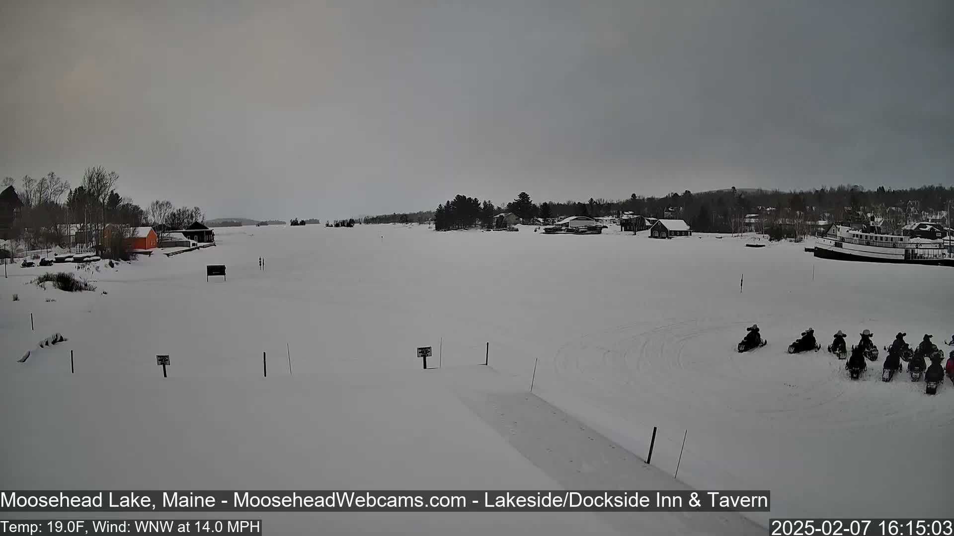A snow-covered lake with a group of snowmobiles gathered on the ice, under an overcast sky.