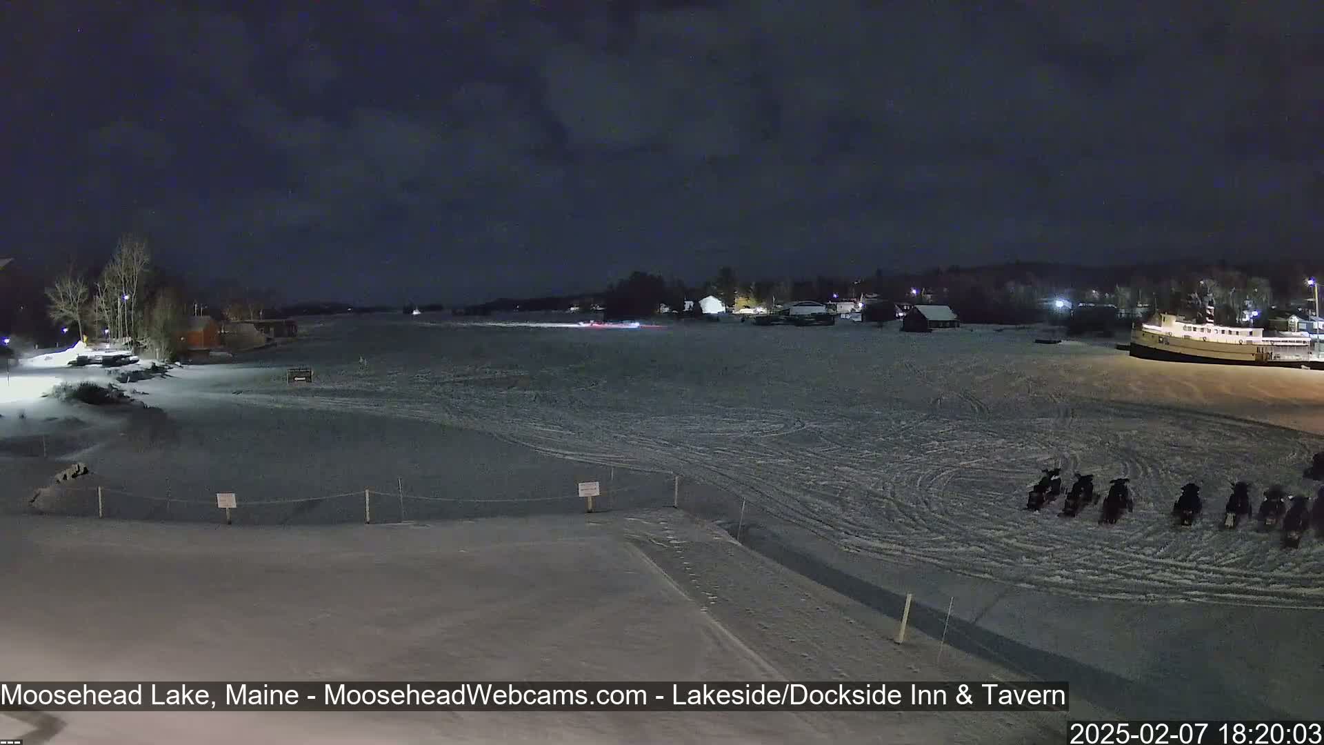 A snow-covered frozen lake at night is viewed from a shoreline, showing a few buildings in the distance and several snowmobiles parked on the ice.