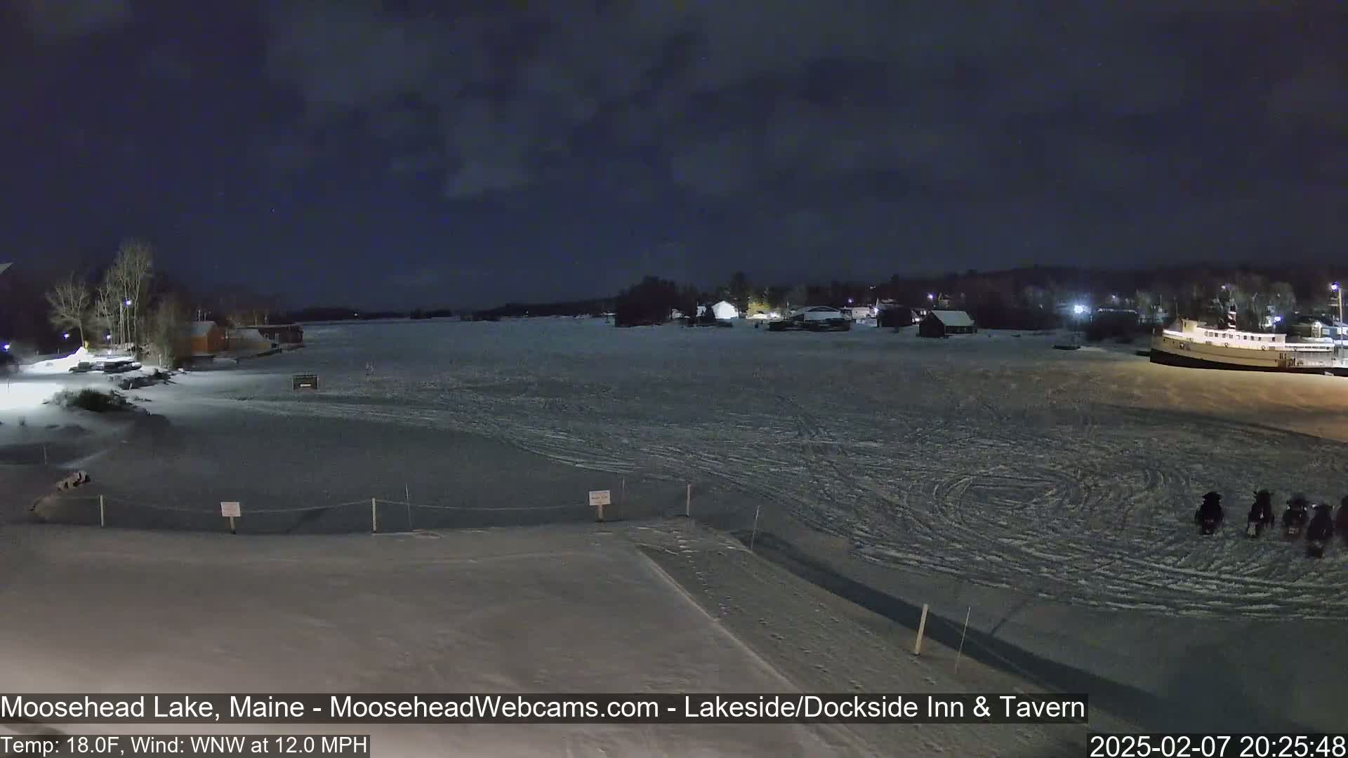 A snow-covered frozen lake at night, with several buildings and a boat visible in the distance, and snowmobiles parked on the ice in the foreground under a dark, cloudy sky.