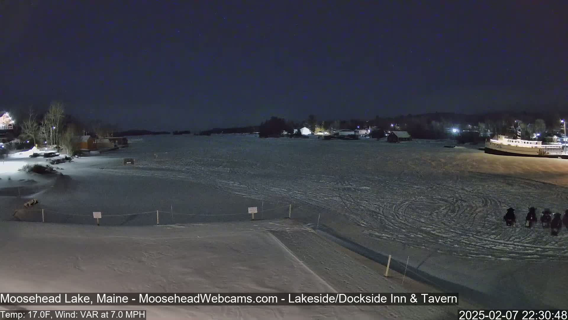 A frozen lake at night, under a dark sky with a few stars, shows a few snowmobiles on the ice near some buildings and a boat.