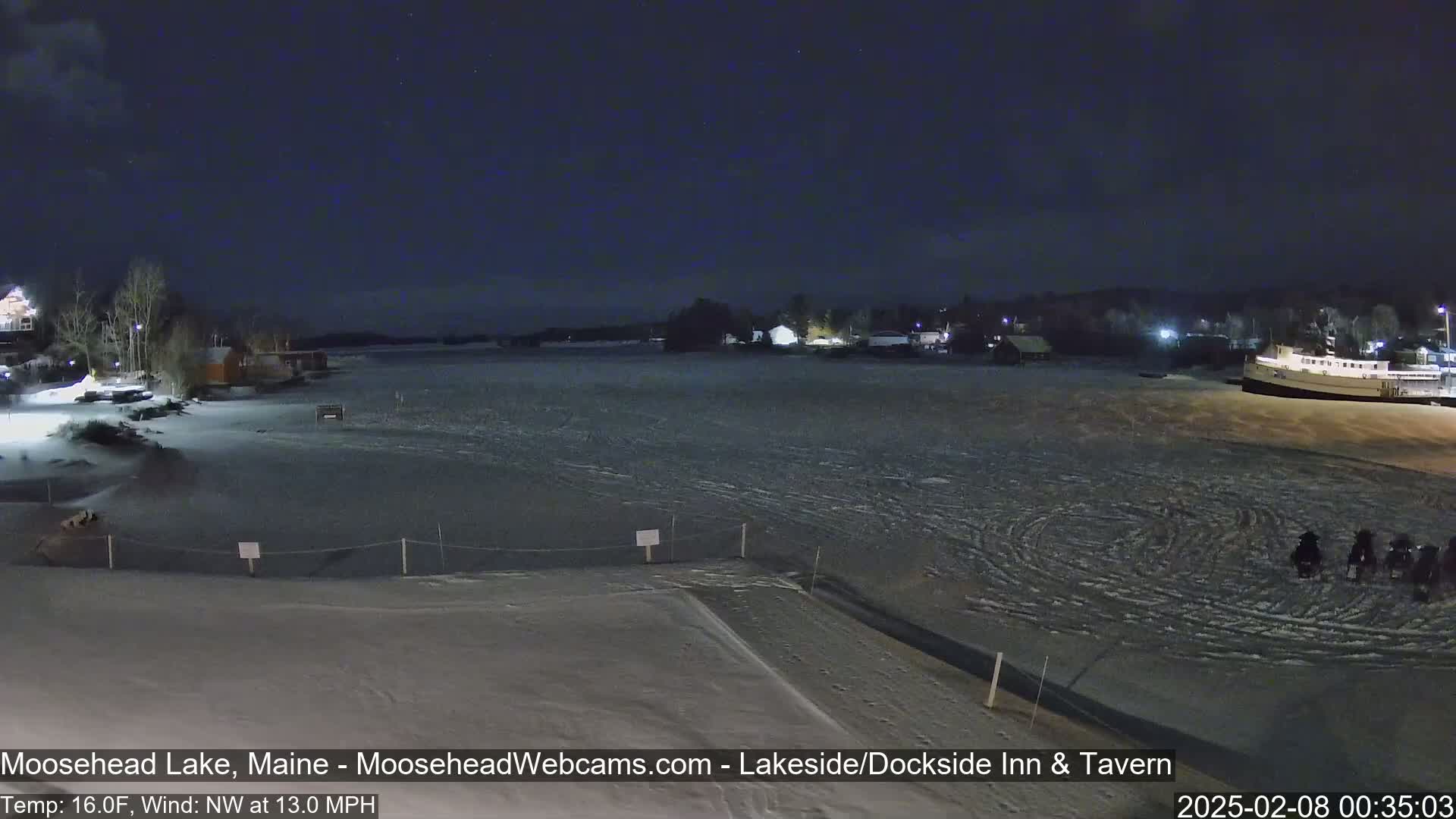 A nighttime view of a snow-covered lake with buildings and trees in the background under a clear, starry sky, with a northwest wind.