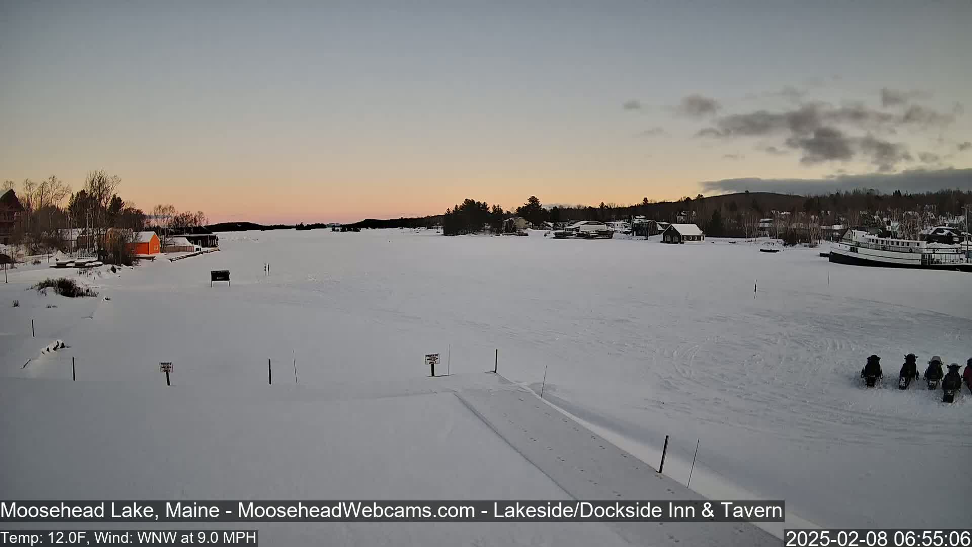 A snow-covered frozen lake at sunset, with several snowmobiles parked on the ice and buildings visible in the distance under a partly cloudy sky.