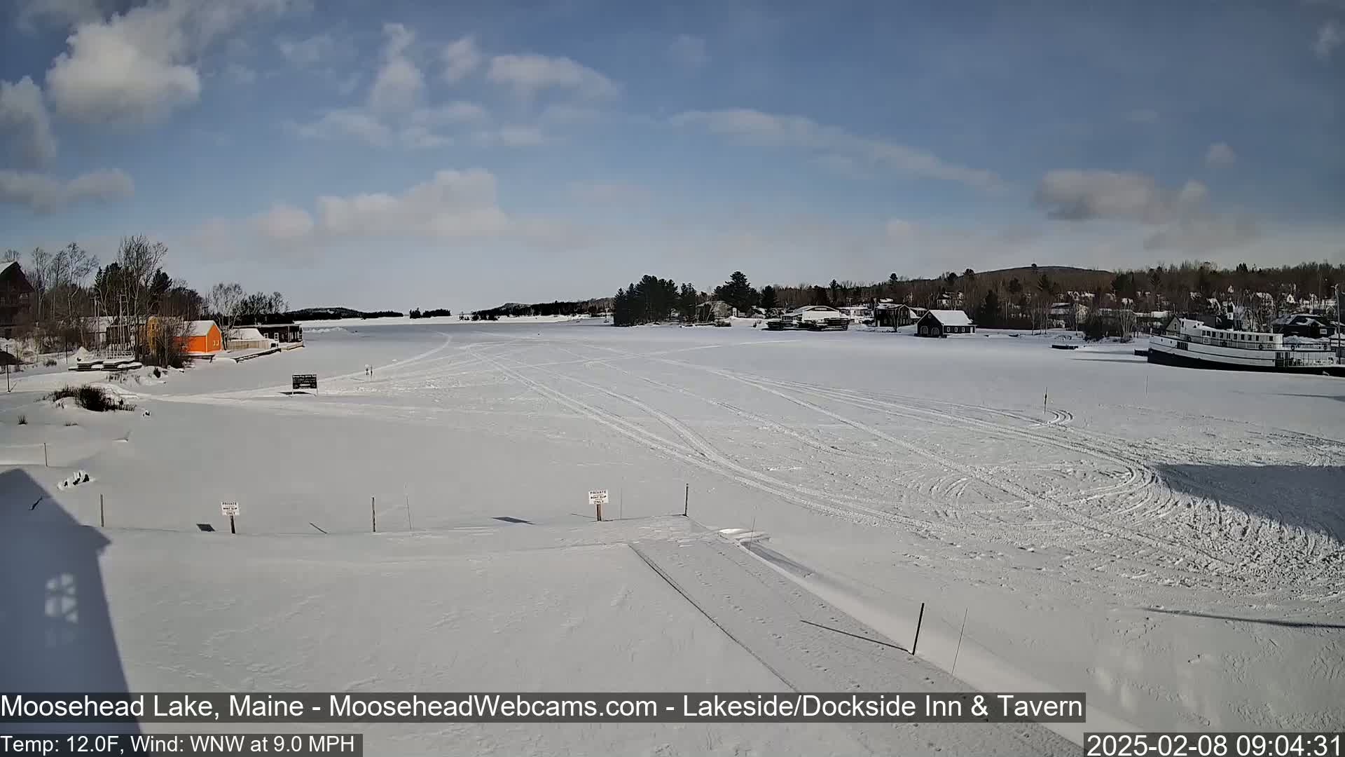 A frozen lake, under a partly cloudy sky, shows tire tracks on the ice, with buildings and a boat visible along the shoreline.
