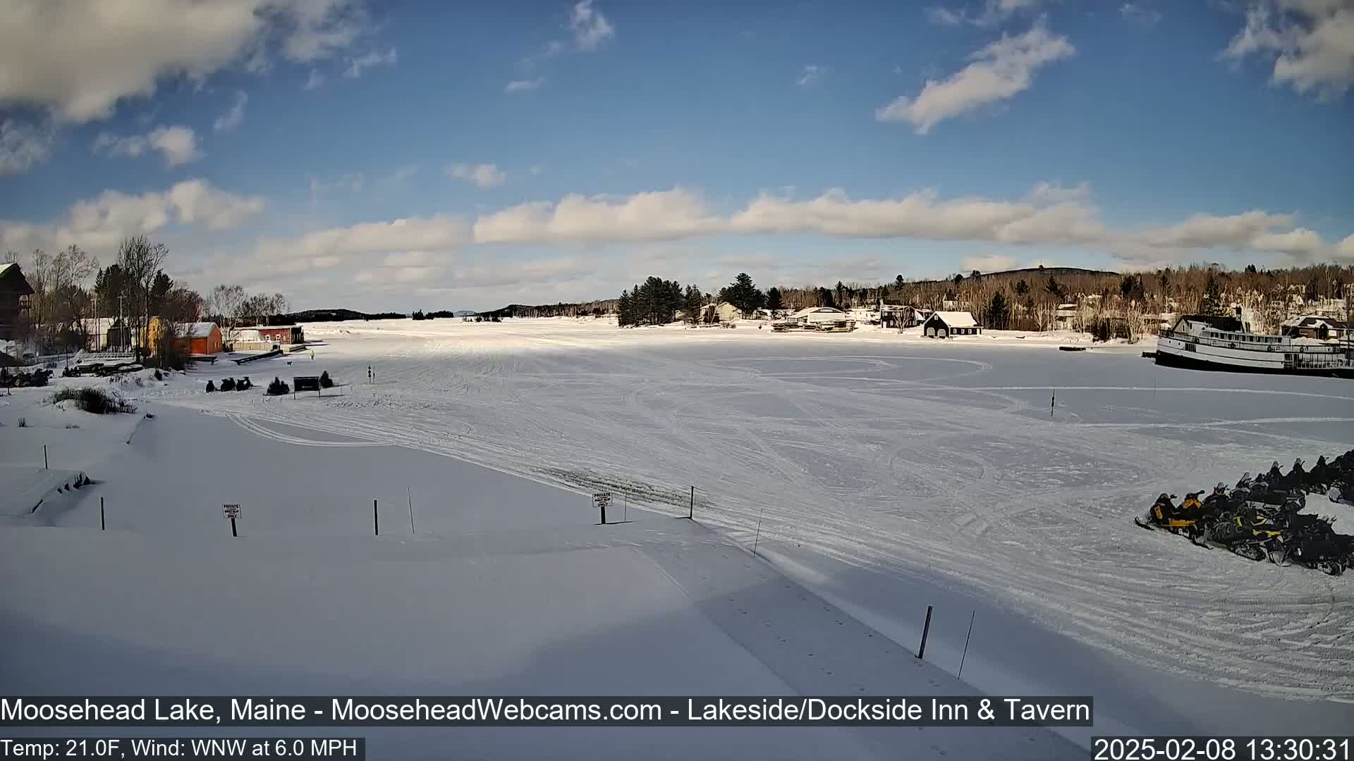 A snow-covered frozen lake with buildings and snowmobiles on the shore under a partly cloudy blue sky.