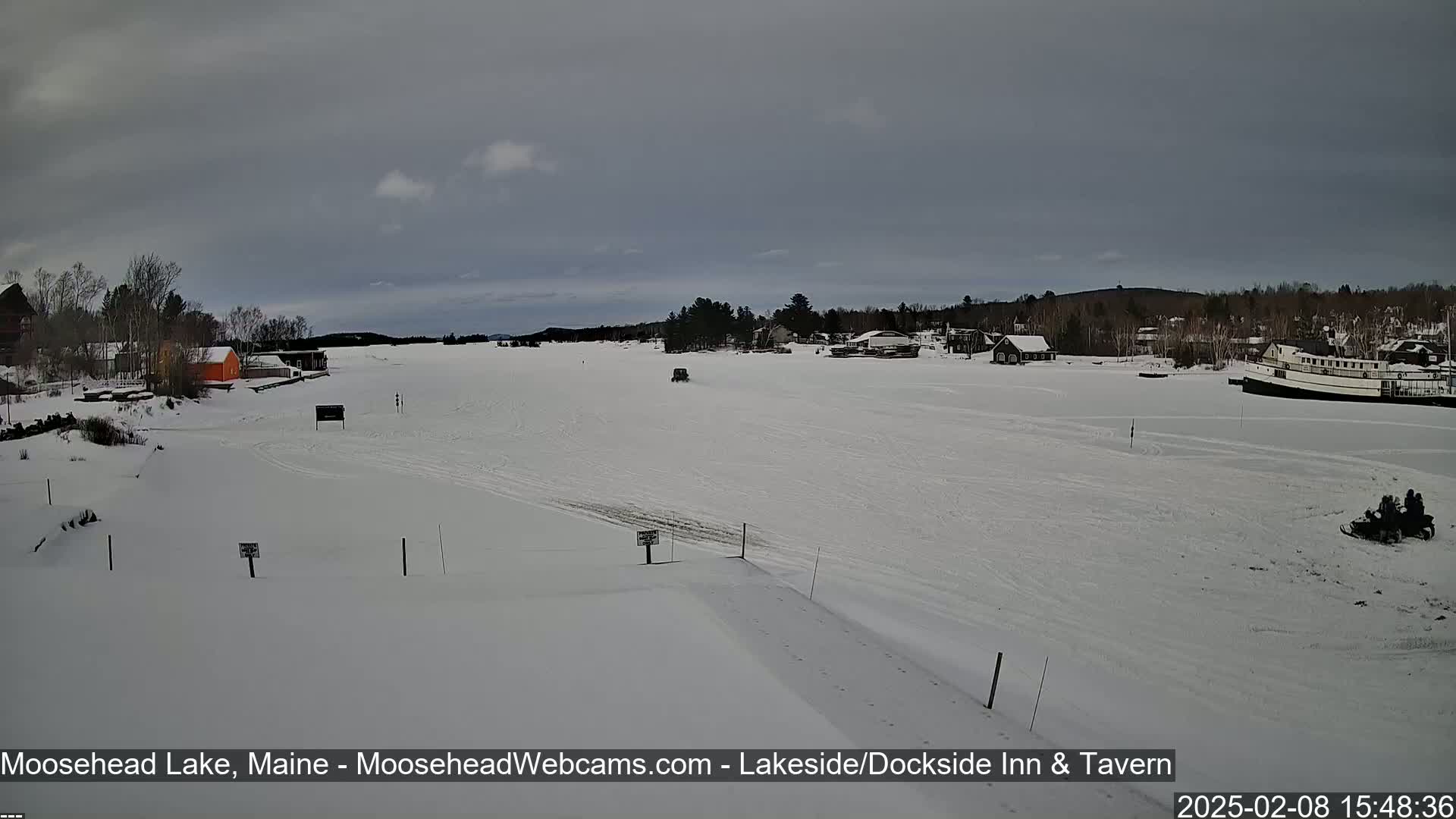 A snow-covered frozen lake is seen with a few buildings and snowmobiles visible in the distance under an overcast sky.