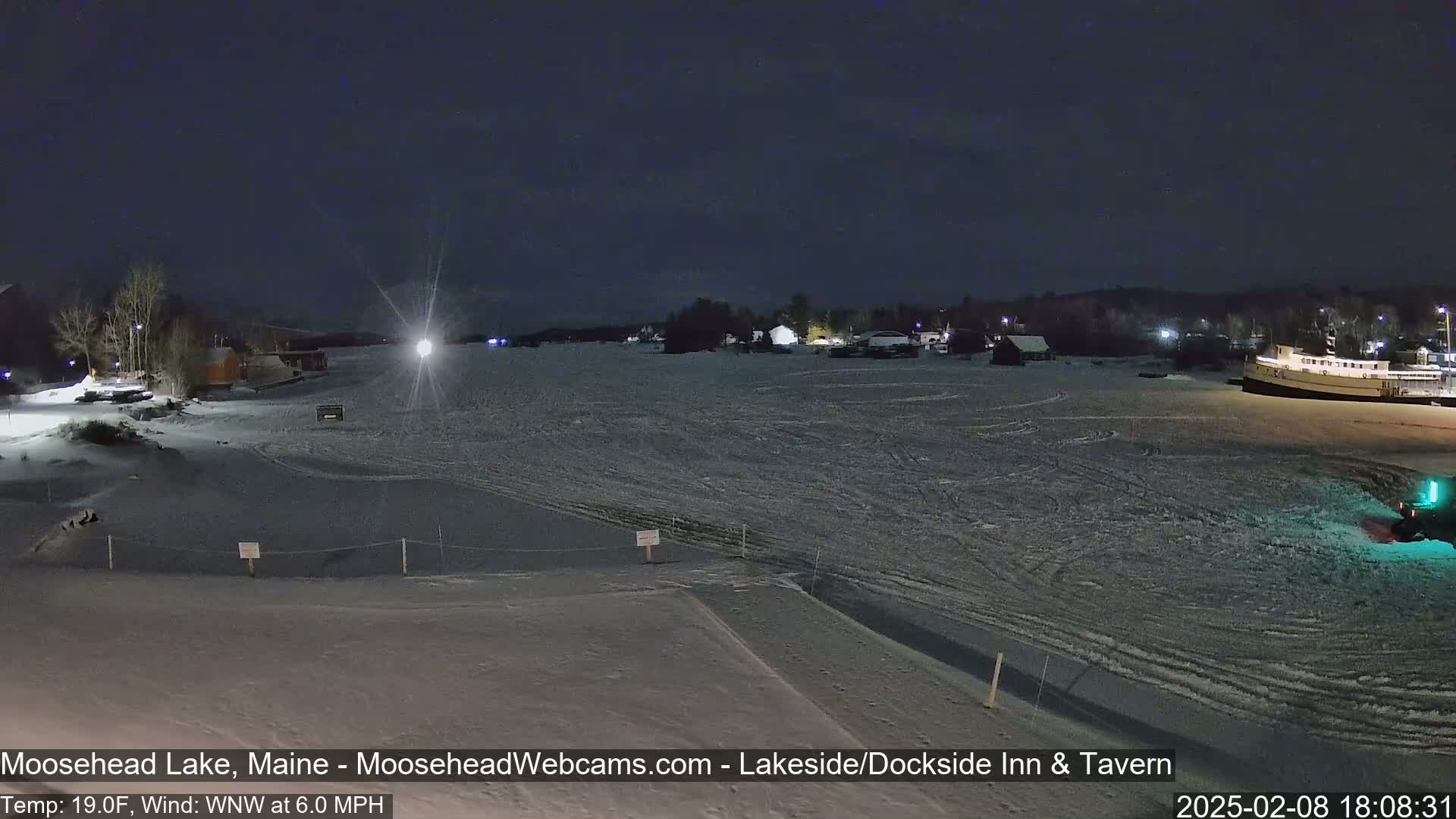 A snow-covered frozen lake at night, with a few buildings and a boat visible in the distance under a dark sky.