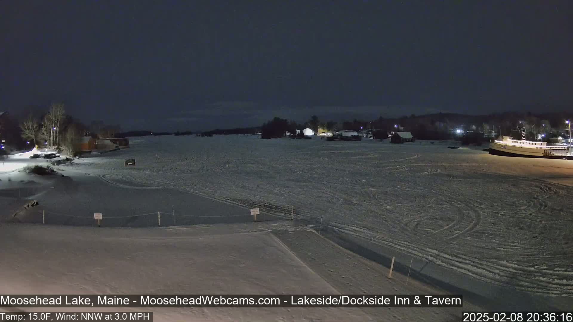 A snow-covered frozen lake at night, with a few buildings and a boat visible in the distance under a dark sky.