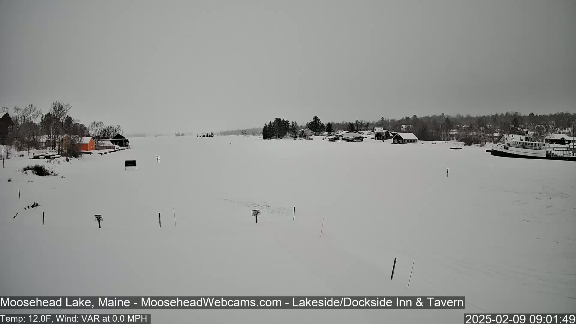 A snow-covered lake with a few buildings and a boat on the far shore under an overcast sky.