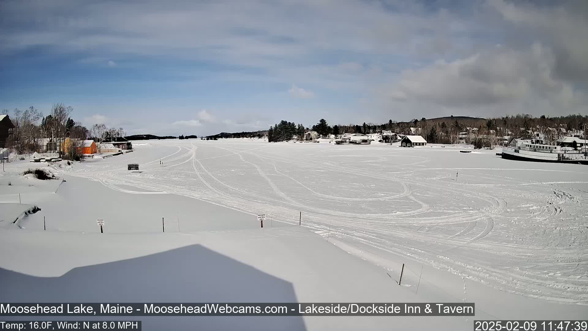 A snow-covered frozen lake is seen with several buildings and a boat in the distance under a partly sunny sky.