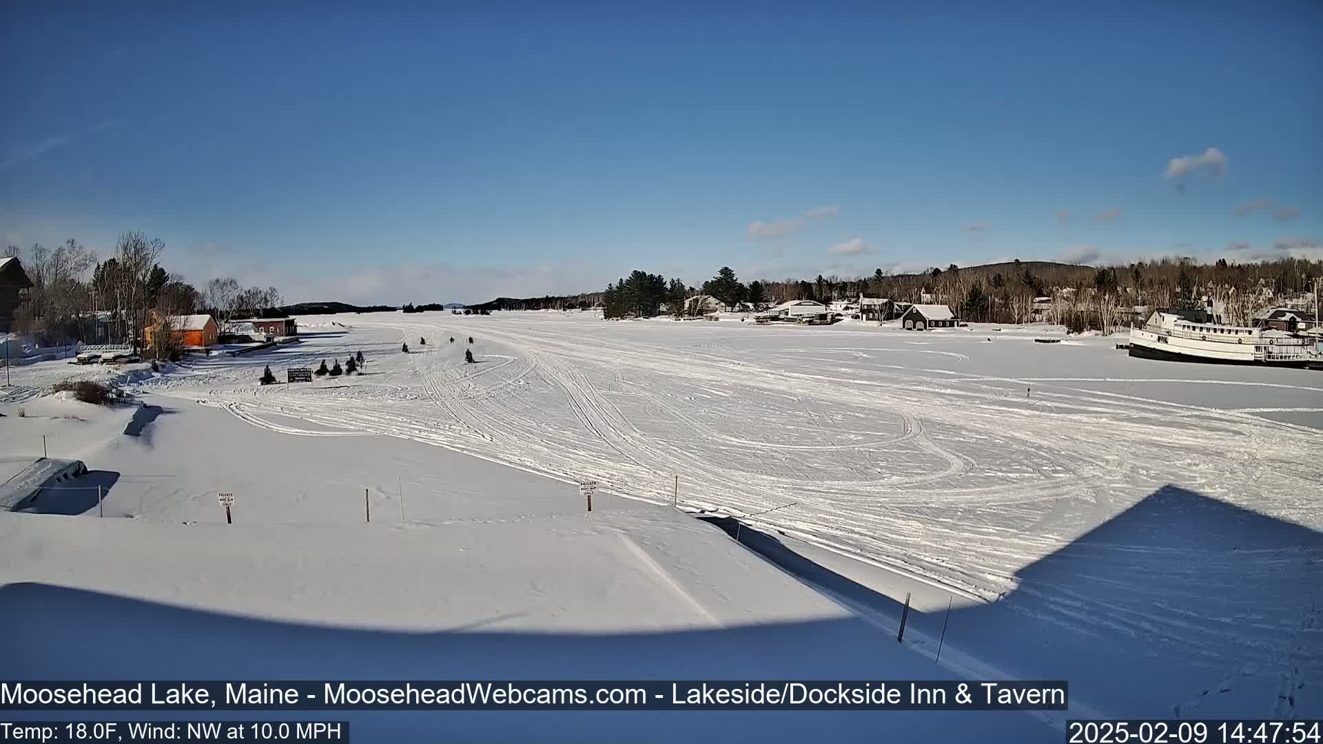 A snow-covered lake with snowmobile tracks, a few buildings, and a large boat are visible under a clear blue sky.